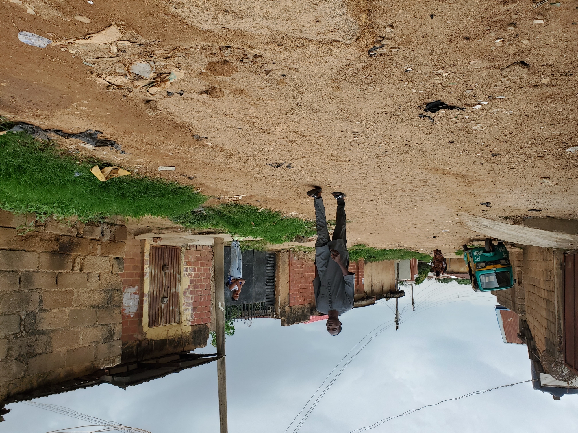 The image shows a dirt road in what appears to be a residential area of Jos, Nigeria. The sky is overcast. Debris is scattered along the road. A man in a gray suit stands in the middle of the road. Another person stands further down the road near a house. A green and yellow auto-rickshaw is parked to the side. Houses made of brick and cement blocks line the road. Power lines are visible overhead.