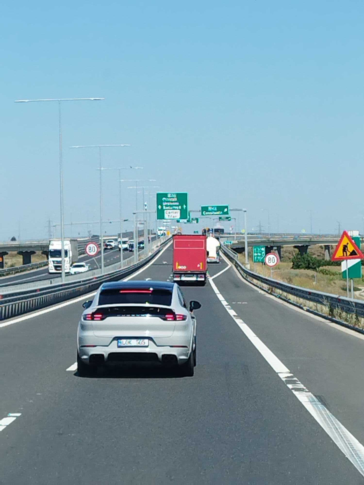 The image depicts a highway scene in the vicinity of Glina, Romania, under a clear blue sky. A silver Porsche Cayenne is prominently featured in the foreground, driving on a multi-lane highway. The vehicle's license plate reads "MLOK 505". Other vehicles, including trucks and cars, are visible further down the road. Traffic signs, including speed limit signs (80) and construction zone warnings, are present along the highway. The environment is characterized by a paved road, guardrails, and vegetation on the sides. The overall impression is that of a typical highway transit.
