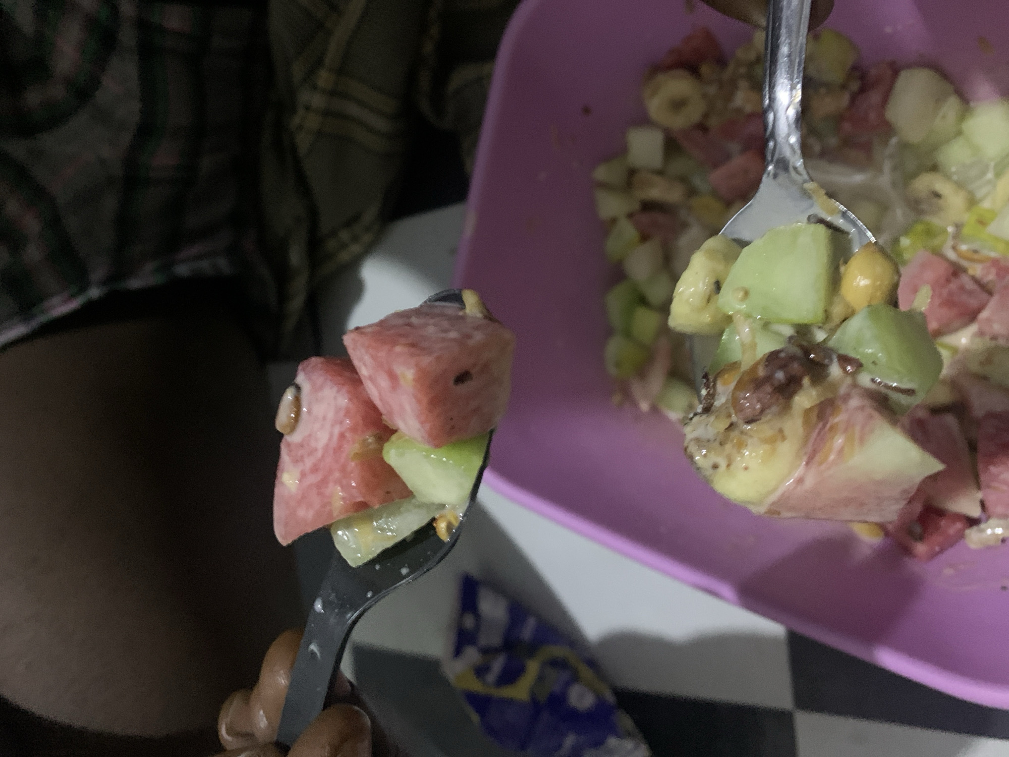 The image captures a close-up perspective of a person eating fruit salad in what appears to be an indoor setting in Rafin Par, Nigeria.
In the immediate foreground, a dark-colored fork, held by a person's dark-skinned hand, is elevated, showcasing a portion of fruit salad. In the mid-ground, a silver spoon is visible, scooping more fruit salad from a vibrant purple bowl.
The fruit salad consists of various diced fruits, including red and pink pieces of watermelon, green chunks that resemble cucumber or green apple, and yellowish-white slices consistent with banana. The fruits are generously coated in a creamy, light-colored dressing or yogurt, with small, darker specks possibly indicating nuts or other inclusions.
The person's arm and torso, covered in a dark plaid-patterned fabric, are partially visible and blurred in the background on the left side of the frame. The surface beneath the bowl and fork displays a distinctive black and white checkered pattern, suggesting a table or floor. A small, blurry object, blue and white in color and possibly a wrapper, rests on this checkered surface near the fork. The overall lighting is dim, indicating that the scene is likely taking place in the evening or at night. No discernible text is visible anywhere in the image. The scene conveys a casual, personal moment of enjoying a fresh, mixed fruit meal.