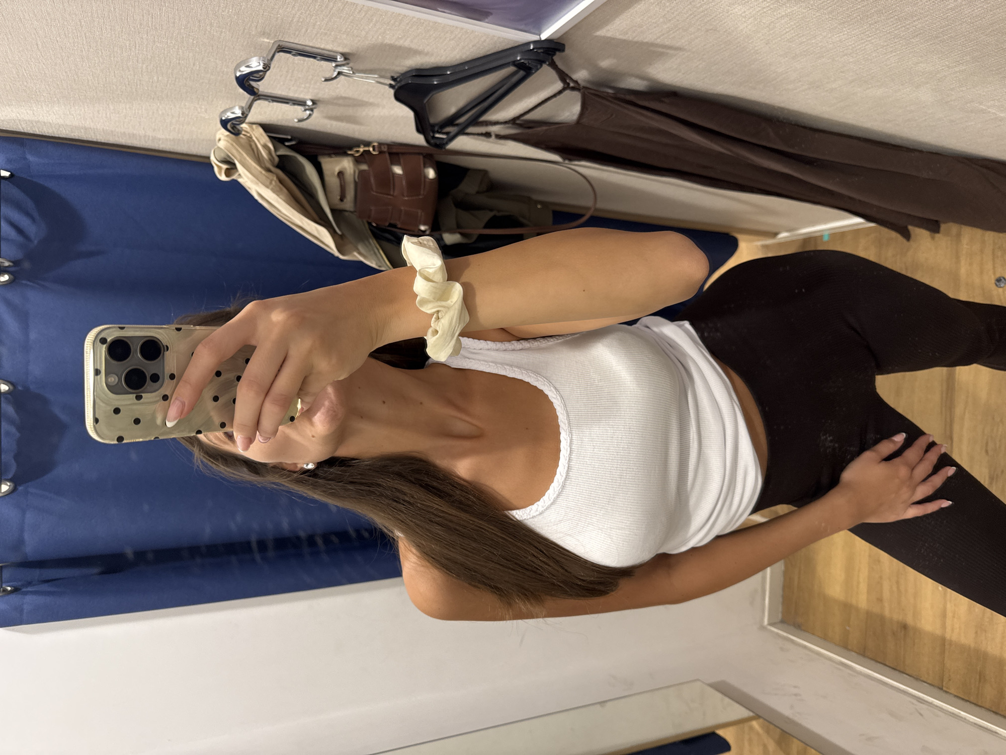 A woman in a white tank top and dark leggings takes a mirror selfie. She is likely in a fitting room, as indicated by the clothes hangers, clothing, and blue curtain in the background. The location cannot be determined as Burgas, Bulgaria based on the image alone.