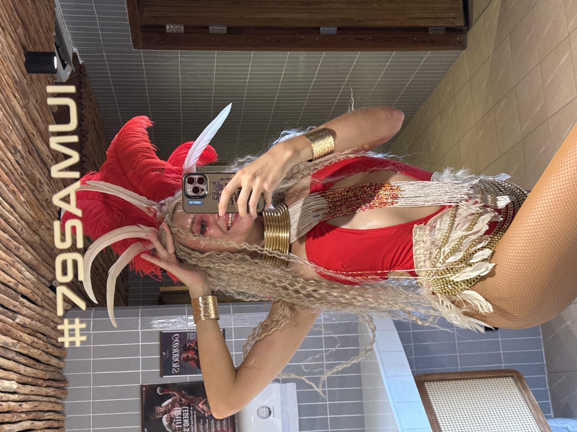 In Ko Samui District, Thailand, a woman with blonde curly hair and elaborate makeup poses for a mirror selfie in what appears to be a restroom. She is wearing a red, one-piece swimsuit adorned with gold chains and white feathers, a large gold neckpiece, gold cuffs on her wrists, and a feathered headpiece with red and white plumes. Her right hand holds a smartphone, capturing the reflection. The background features grey tiled walls, a wooden ceiling, a dark wooden door, and a wooden-framed chair with a woven seat. In the reflection, a hashtag '#795AMUI' is visible above her head.
