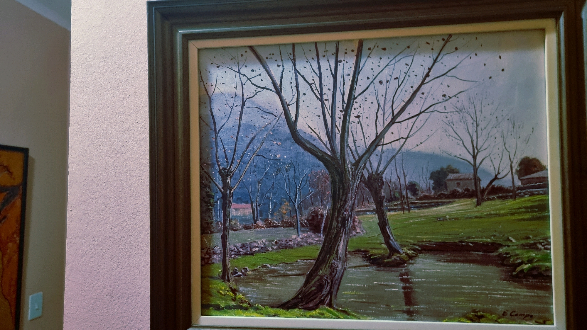 The image shows a framed oil painting of a rural landscape. The painting depicts a body of water flowing through a grassy area with bare trees. In the background, there are houses and hills. The painting is signed "E. Campo". The location is specified as Sant Joan Les Fonts, Spain. The painting is hanging on a textured, light pink wall, with a portion of another framed artwork visible to the left. The painting itself is framed with a dark wooden outer frame and a lighter inner frame.
