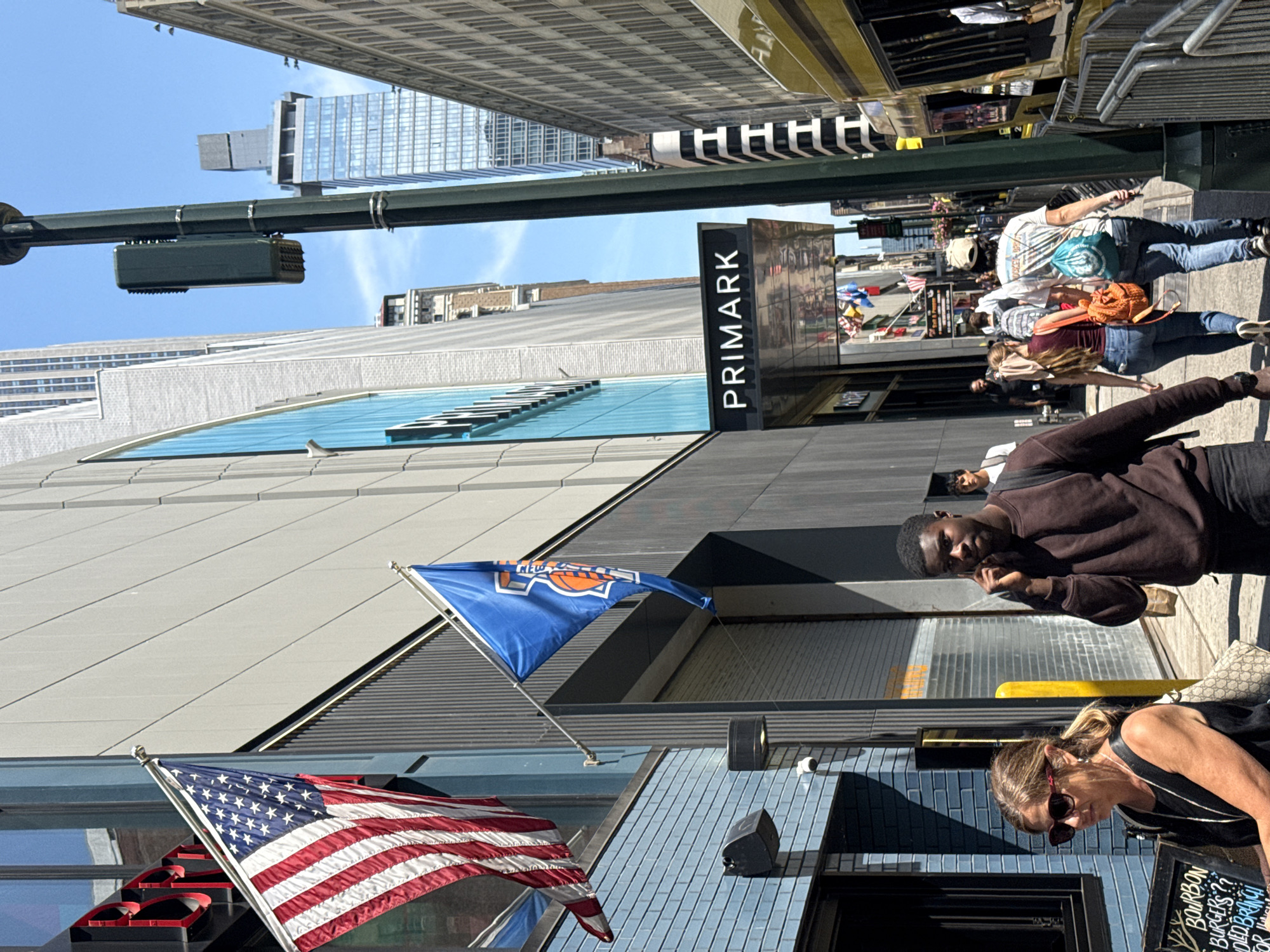 This is a vertical, outdoor photograph taken on a sunny day in a bustling urban environment, likely New York City. The scene is dominated by modern architecture with large glass windows and a prominent "PRIMARK" sign. Several people are visible, some walking, others looking at their phones.
In the foreground, an American flag waves on a flagpole, juxtaposed with a blue flag featuring the New York Knicks logo. A man in a brown sweatshirt is looking towards the camera, while a woman with blonde hair and sunglasses is examining a sign.
The background shows tall buildings with glass facades and metal accents, suggesting a commercial district. The sunlight casts distinct shadows, indicating it's likely midday. The overall atmosphere appears to be one of everyday urban activity, with a mix of shoppers and pedestrians.
Visible text includes "PRIMARK" on a storefront sign and some text on a chalkboard sign which is partially obscured but appears to advertise "BOURBON BURGERS?". The image captures a slice of life in a vibrant city.
