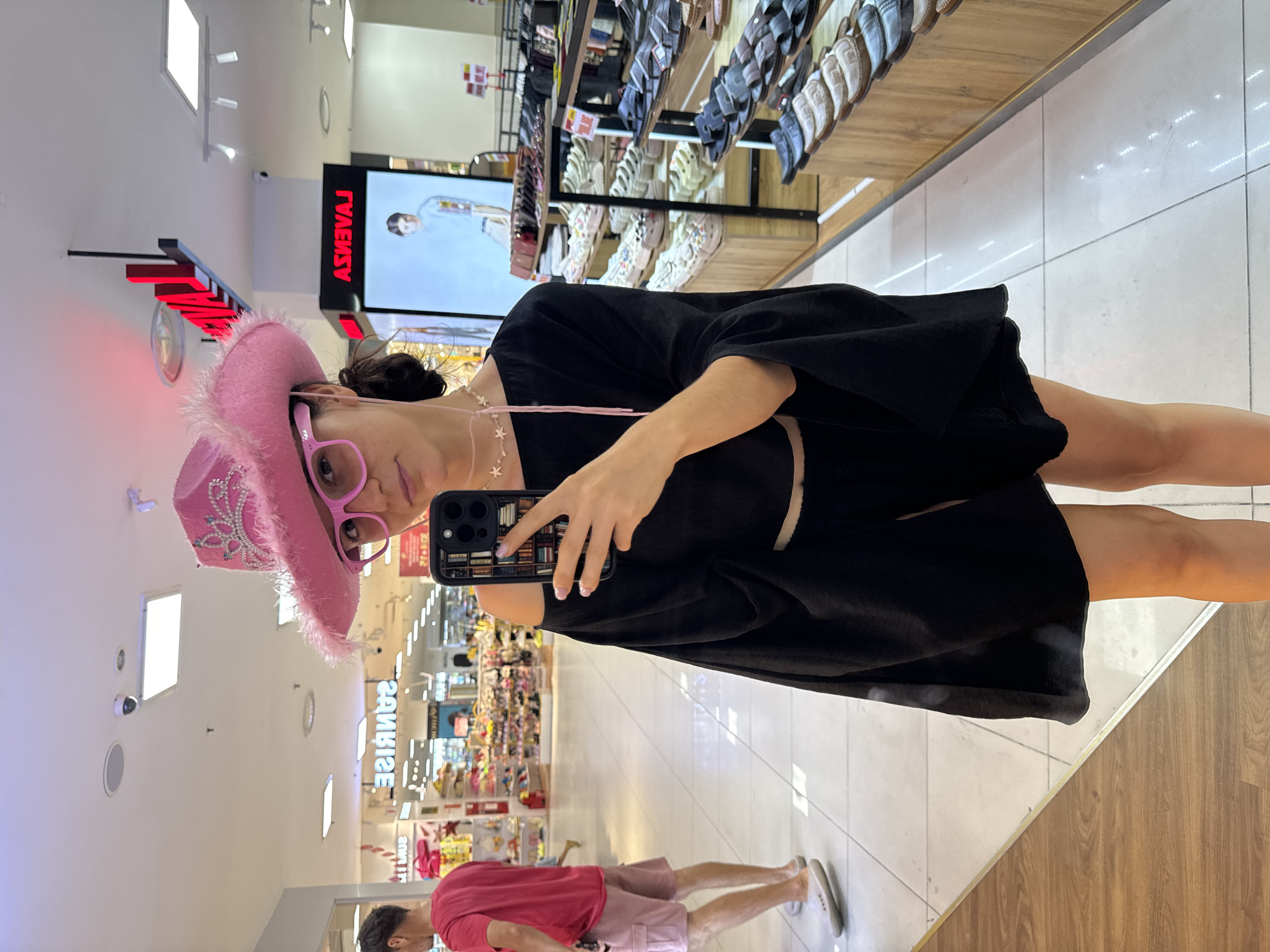 A woman in a black dress, pink cowboy hat, and pink glasses takes a selfie with a black phone. The scene is inside a store, likely a shopping mall or department store, with shelves displaying shoes in the background. Another person in a red shirt is visible in the lower part of the image. Based on the "Sunrise" sign, this could be Phú Thủy, Vietnam. The woman is positioned near a display of footwear, and the environment suggests a retail setting.