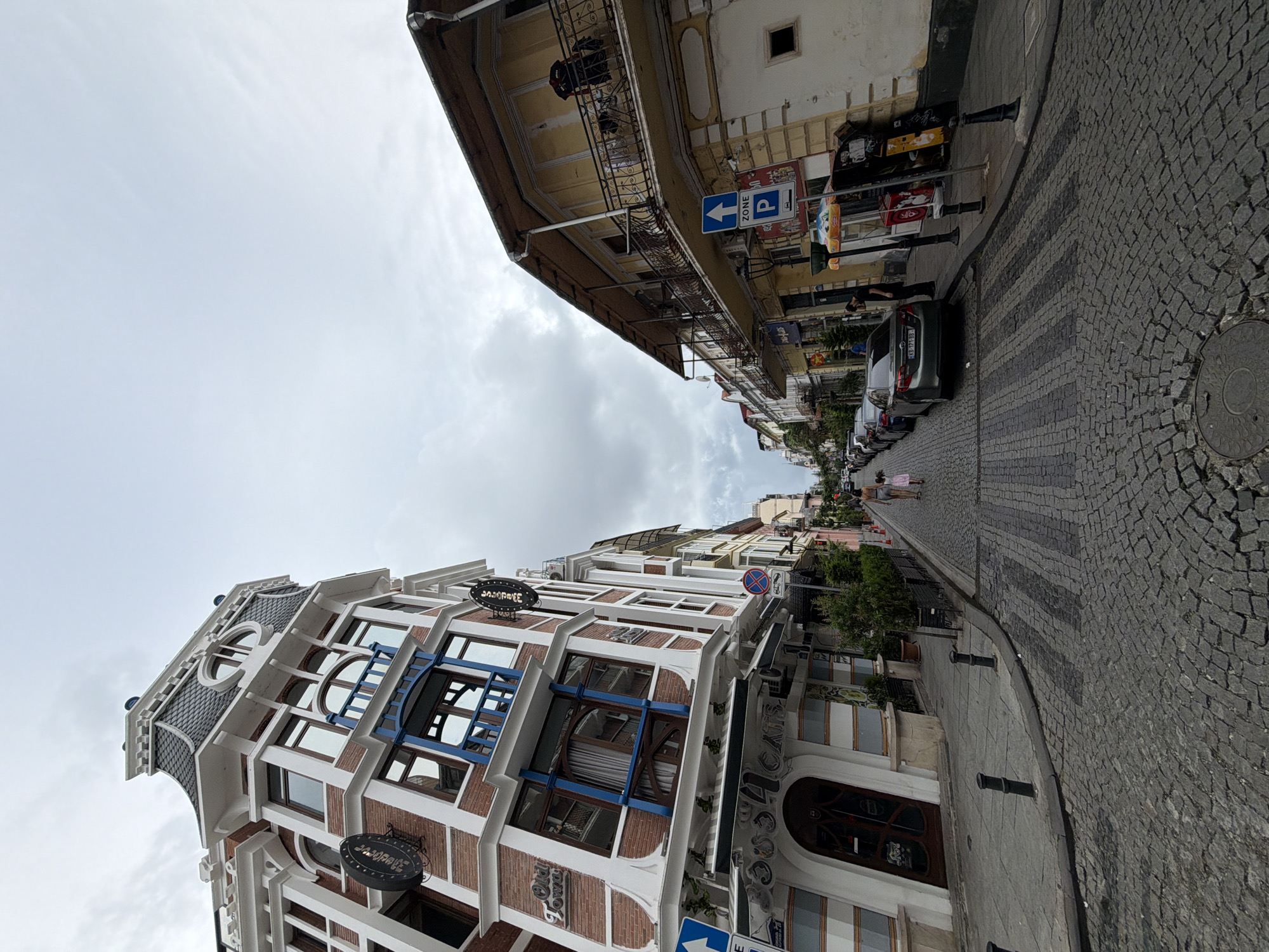 A street scene in Batumi, Georgia, features a cobblestone road lined with buildings. On the left, a multi-story building with a distinctive mansard roof and brick facade has ornate window frames and a prominent sign for "Old Town" and "4 Cats". Awnings extend over the ground-floor businesses. On the right, a building with a yellow facade and balconies overlooks the street. Several cars are parked along the side of the road, and a few people are visible walking on the sidewalk. The sky is overcast with gray clouds.
