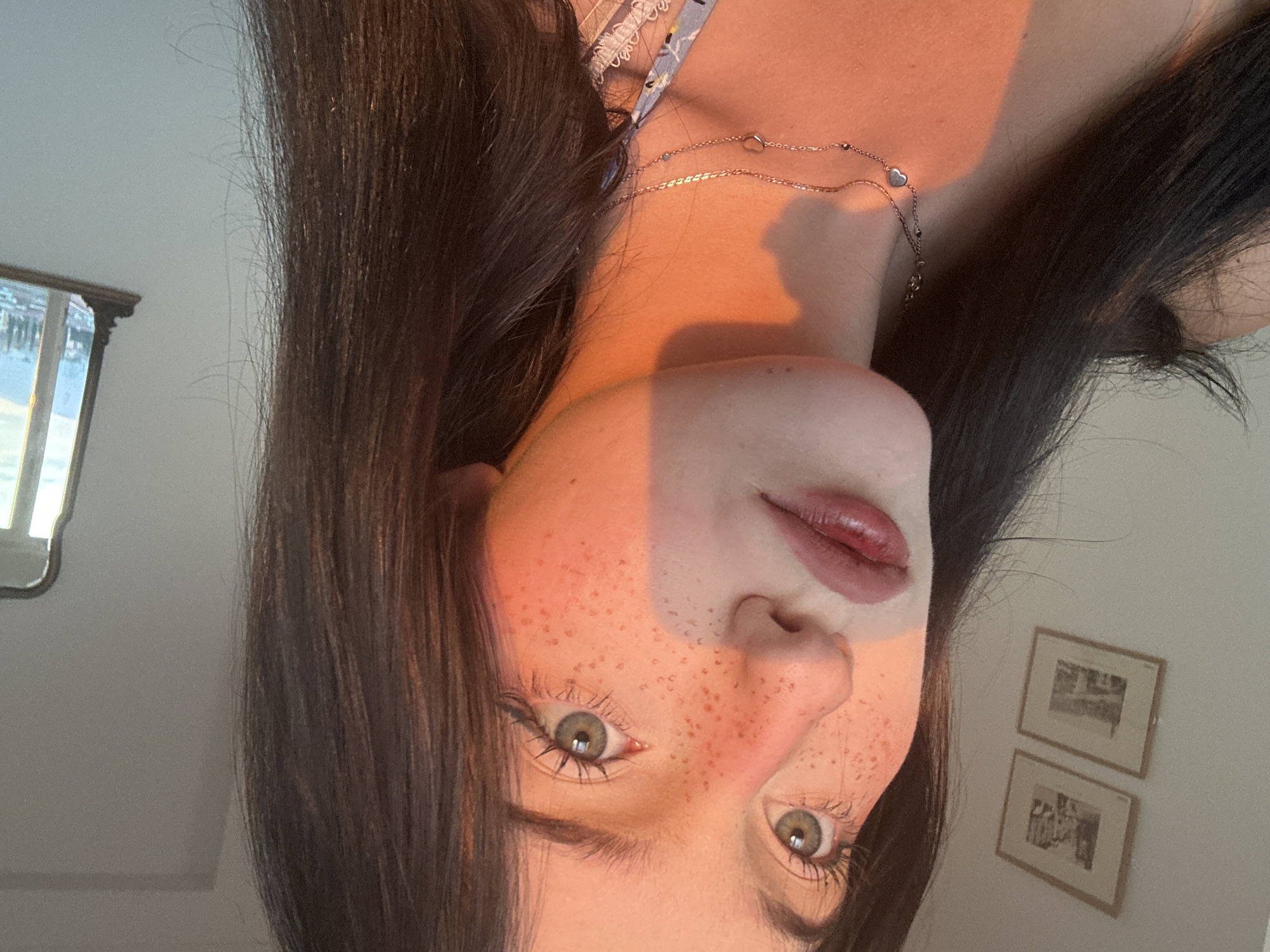 A close-up portrait of a young woman with dark hair and fair skin, adorned with freckles and a light pink lipstick. The warm glow of sunset illuminates her face, casting a soft orange hue and a distinct shadow across her nose and cheek. She is wearing a delicate silver necklace with a heart pendant. In the background, a white wall is decorated with two framed pictures. To the right, a decorative mirror reflects a view of a city and mountains, possibly Verona, Italy.