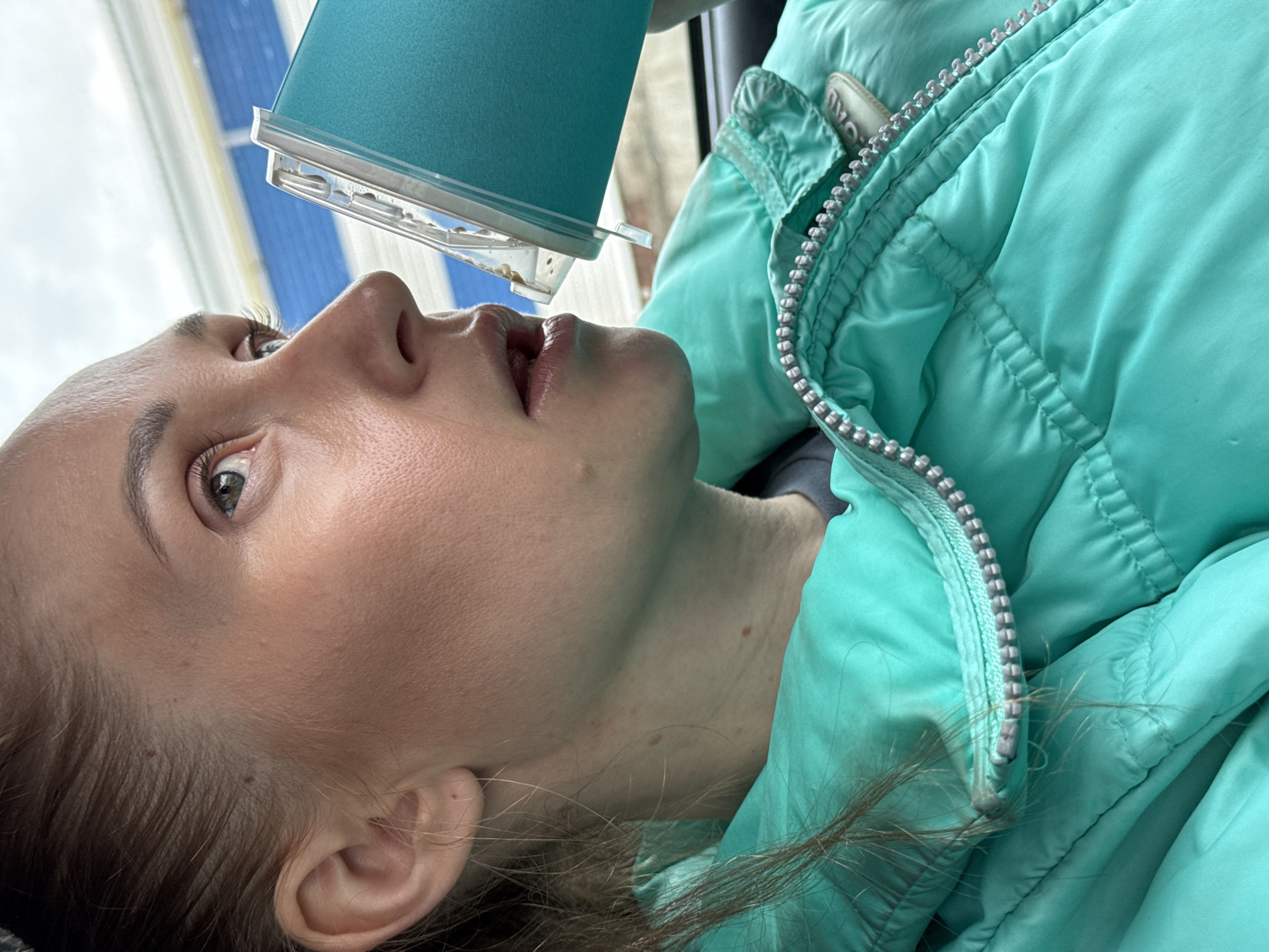 A Caucasian woman lies supine, with a small blue rectangular container held above her mouth, appearing to dispense small, white objects. She is wearing a light blue puffer jacket. The background shows a blurry view of a building with blue and white horizontal stripes, possibly seen through a window. The location cannot be determined to be Chaikovsky, Russia, based on the image.