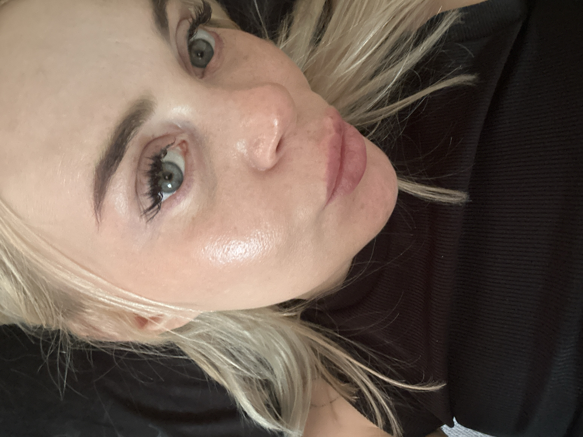 A close-up portrait of an adult female with blonde hair, looking directly at the camera. Her eyes are light blue-grey, framed by prominent dark eyelashes. Her eyebrows are dark and defined. Her fair skin exhibits a noticeable sheen or highlight on her right cheekbone and nose. Minimal skin texture is visible on her forehead. Her lips are medium-full, with a pink/mauve hue, and are slightly pursed. She is wearing a dark, ribbed fabric top. The background is uniformly dark, indistinct, and appears to be fabric, offering no specific environmental context. The location is indoors, in the city of Unknown, Unknown, as no discernible geographical features or unique environmental markers are present.