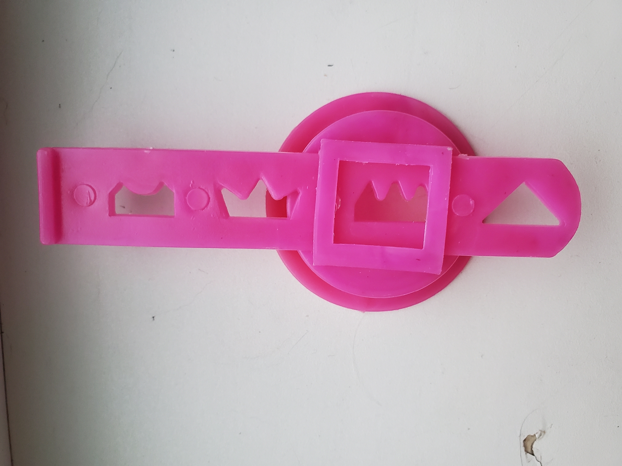 The image shows a bright pink plastic toy or tool with various cutouts and shapes. It is lying on a textured, off-white surface, possibly a windowsill or a wall. The toy has a long, rectangular section with rounded ends at the top, featuring two circular indentations and three rectangular cutouts with jagged edges. Below this section, there is a circular disc. Mounted on the disc is a smaller square frame, also with jagged edges inside. The bottom part of the toy is a wider section with a triangular cutout. The surface it rests on appears to be slightly weathered, with a small area of damage visible in the lower left corner. The lighting suggests an indoor environment with diffused natural light. There are no people, animals, or any other objects present in the image besides the pink plastic item and the surface it is on. The scene is static and appears to be a close-up, overhead shot of the object. This image does not provide any information or context related to the city of Tiraspol, Moldova, or any specific location within it.