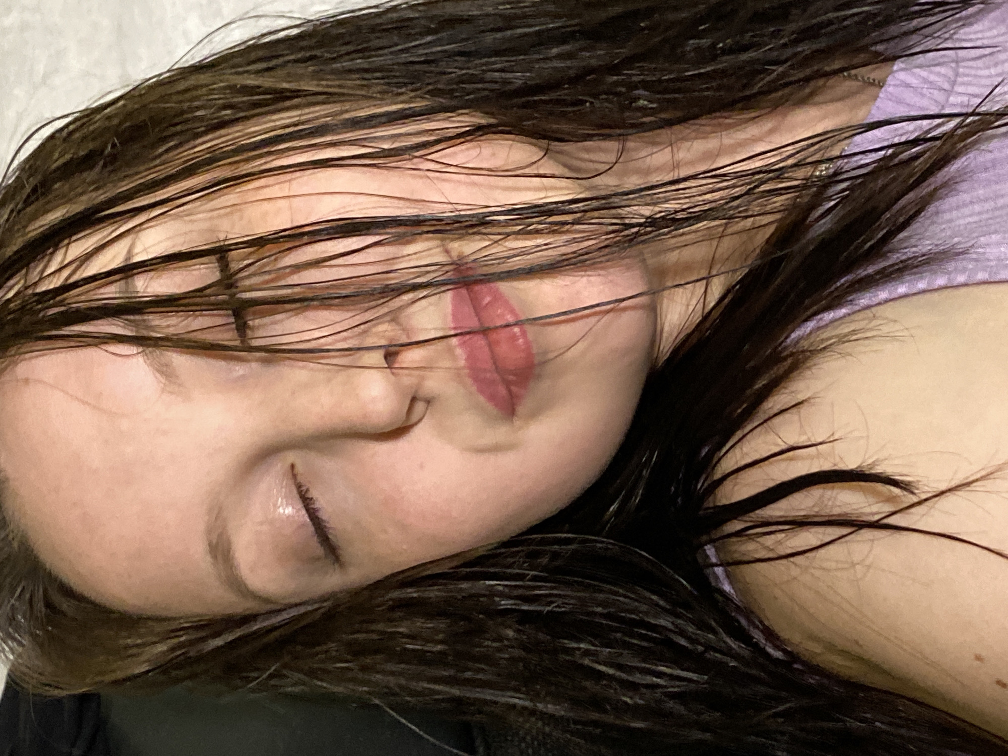The image depicts a person lying down with wet, dark hair spread across the face and surrounding surface. The person's eyes are closed. The face is visible, showing a nose, mouth with pink lipstick, and pale skin tone. The person appears to be wearing a light purple shirt. The location in the city of Chaikovsky, Russia cannot be determined from the image.