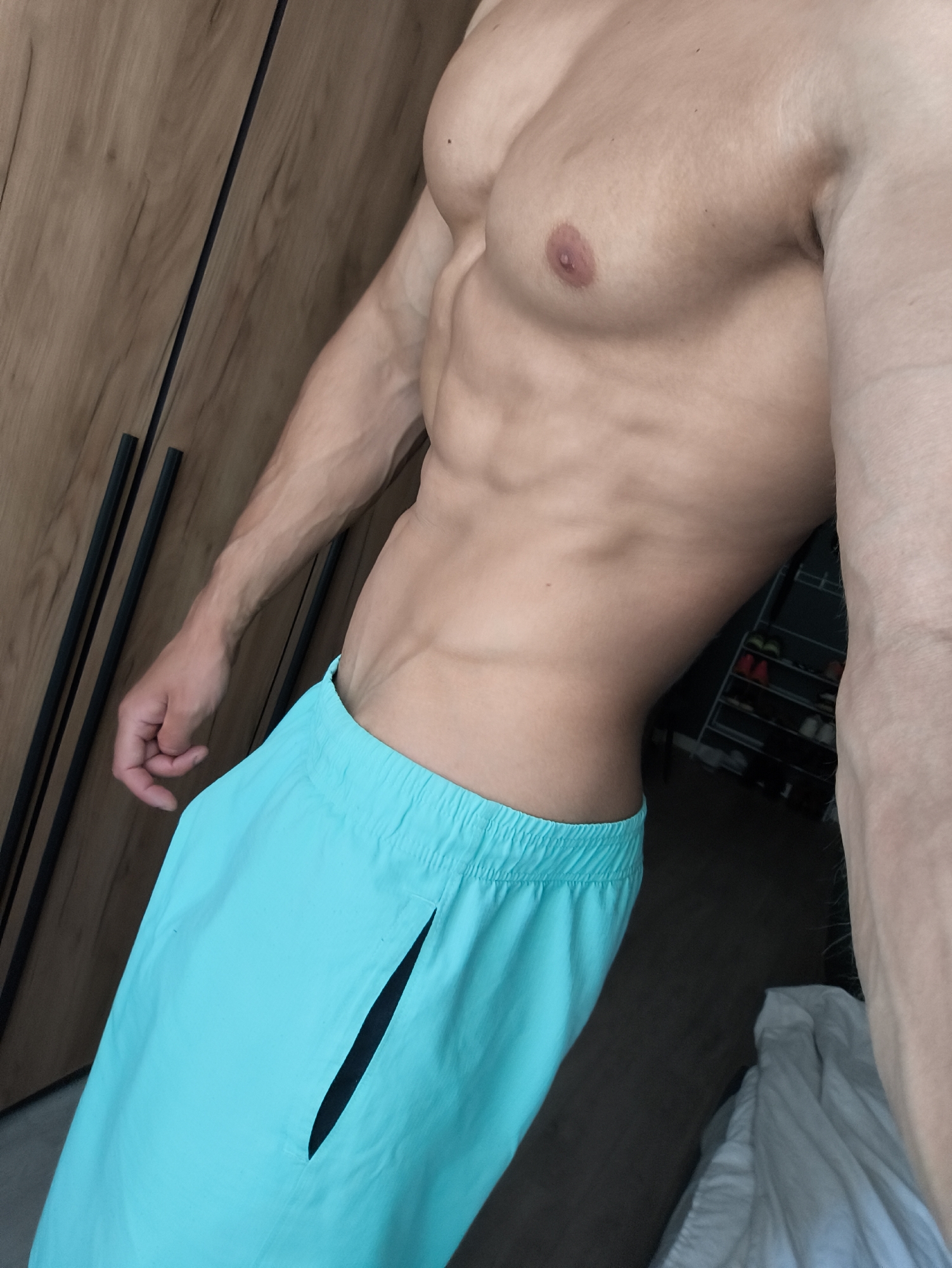 A partial, close-up view of a fit man's torso and legs is presented. The man is shirtless, revealing well-defined pectoral muscles and abdominal muscles. He is wearing bright turquoise shorts with a black slit or pocket detail on the side. The background includes a light brown wood-paneled wall with black handles and a partial view of a room with shelving. There is no evidence to connect the photo to the city of Moscow, Russia.