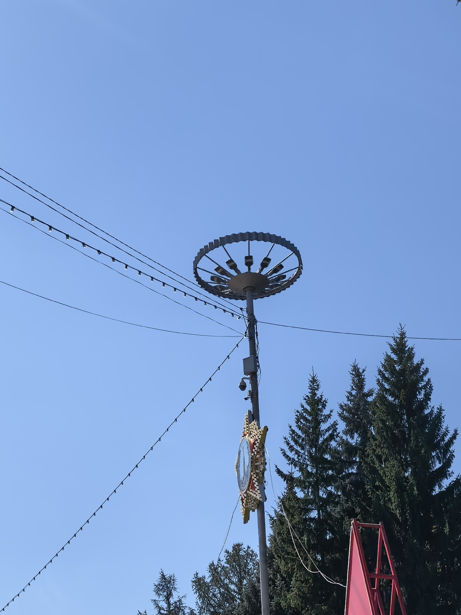 The image, taken on a clear, blue sky day, captures a tall lamppost with a circular, wheel-like fixture at its apex, equipped with multiple lights. Below this, a clock with a decorative, star-shaped frame is mounted on the lamppost. Several strings of small lights are strung across the sky, extending from the lamppost. In the background, the tops of several evergreen trees are visible, and a portion of a red structure, possibly a stand or frame, is present in the lower right corner. This scene appears to be located in the city of Sterlitamak, Russia.