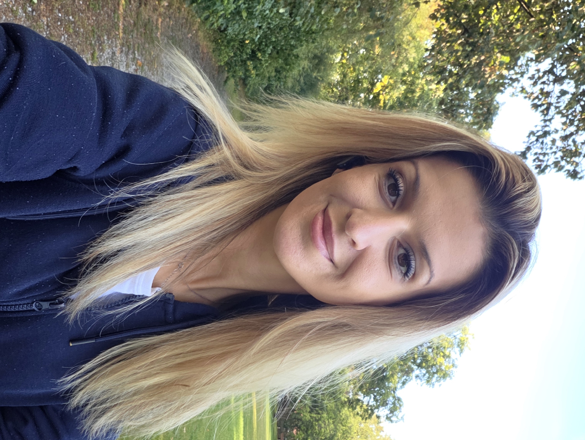 A selfie of a woman with blonde hair is taken outdoors. She is wearing a dark blue hooded sweatshirt over a white t-shirt and a silver necklace. The background shows trees, a path, and a grassy area, suggesting a park or a wooded area. The lighting and background suggest it is daytime. The image does not contain any information that would indicate the location in the city of Munich, Germany.