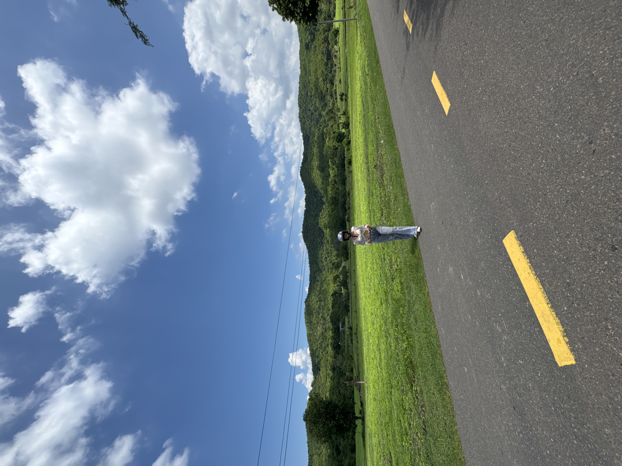 A person wearing a white top, blue jeans, and a helmet stands on an asphalt road with yellow lane markings in Ham Thuan Bac, Vietnam. The road curves slightly to the right. To the left of the road is a vibrant green field. In the background, rolling hills covered in lush green foliage rise, and a clear blue sky with scattered white clouds stretches above. Power lines are visible across the horizon.