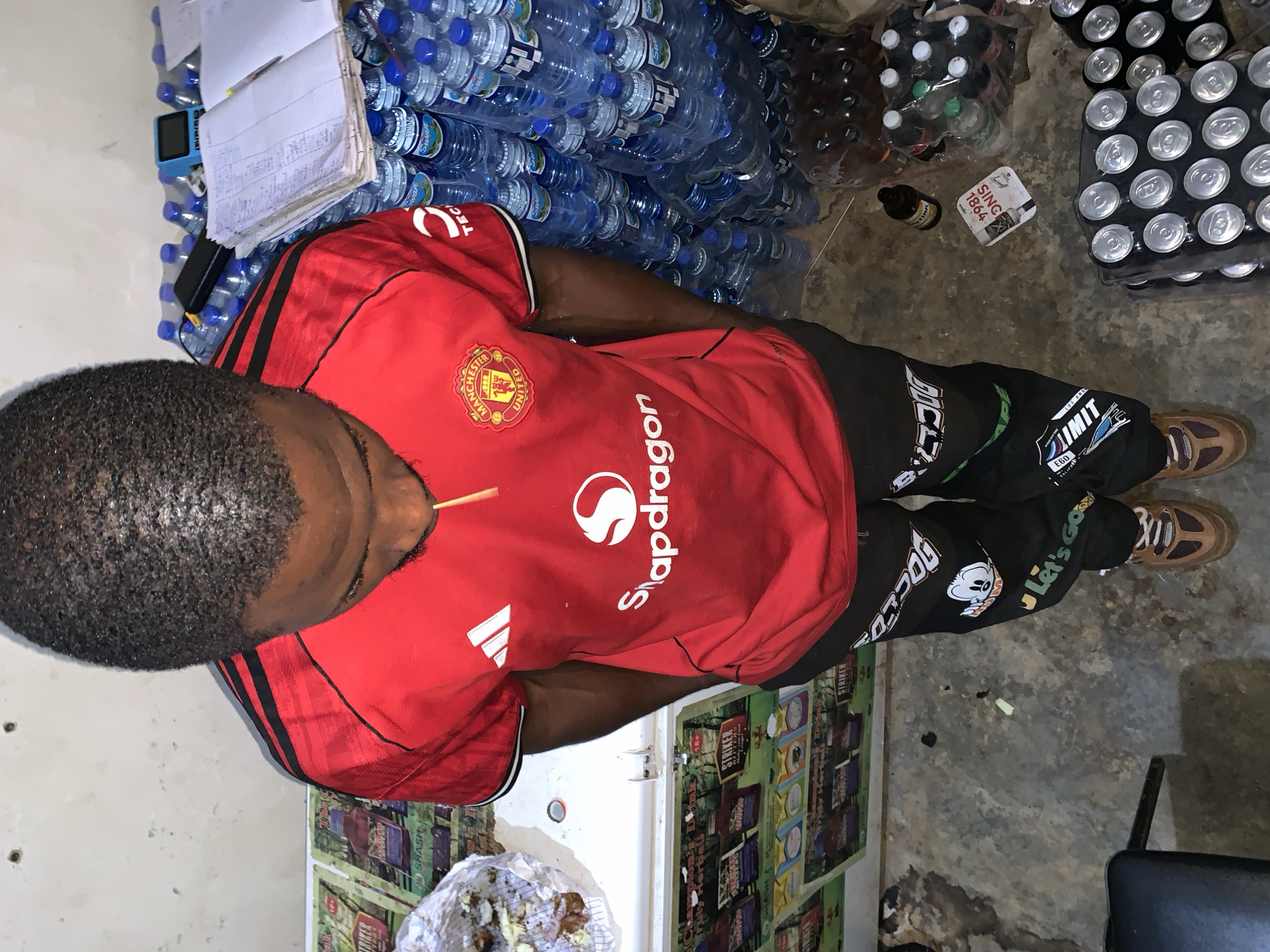 The image depicts a man standing indoors amidst a stock of beverages. He is wearing a red Manchester United jersey with "Snapdragon" printed on it, paired with black pants featuring various prints and brown shoes. The immediate environment includes stacked plastic bottles of water, cans of beverages, and glass bottles of alcoholic drinks. The floor is bare and appears to be concrete. In the background, there's a white refrigerator plastered with "STRIKER" advertisements, and a notebook lying on top of something, possibly a scale, near more bottles of water. A toothpick is visibly sticking out of the man's mouth. The man's location in the city of New Stadium, Nigeria, cannot be confirmed from this image.