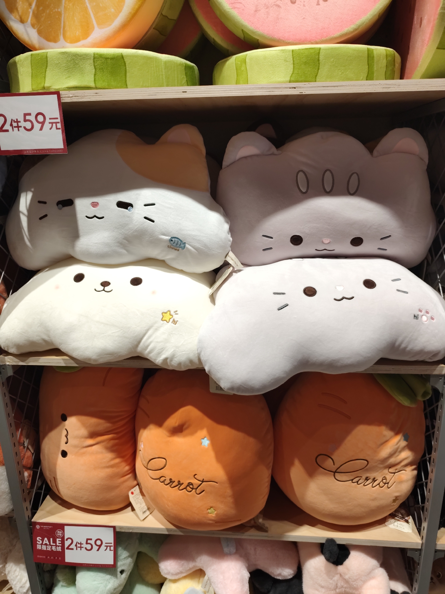 The image shows a retail display of stuffed animal pillows on wooden shelves. The pillows are arranged in stacks. On the top shelf, there are large, round pillows designed to look like slices of fruit, specifically oranges and watermelons. The middle shelf holds four cloud-shaped pillows with embroidered animal faces. Two are white with orange markings and brown eyes, and two are light grey with darker grey eyes and ears. The bottom shelf features two large, orange carrot-shaped pillows with green tops. Below the shelves, there are more plush toys visible, in various colors. A red and white sale sign in Chinese characters is attached to the front of the bottom shelf, advertising "2 items for 59 yuan." The overall scene is a retail environment, likely a toy or home goods store. The city of Daqing, China is mentioned in the prompt but no visual cues in the image confirm this specific location.
