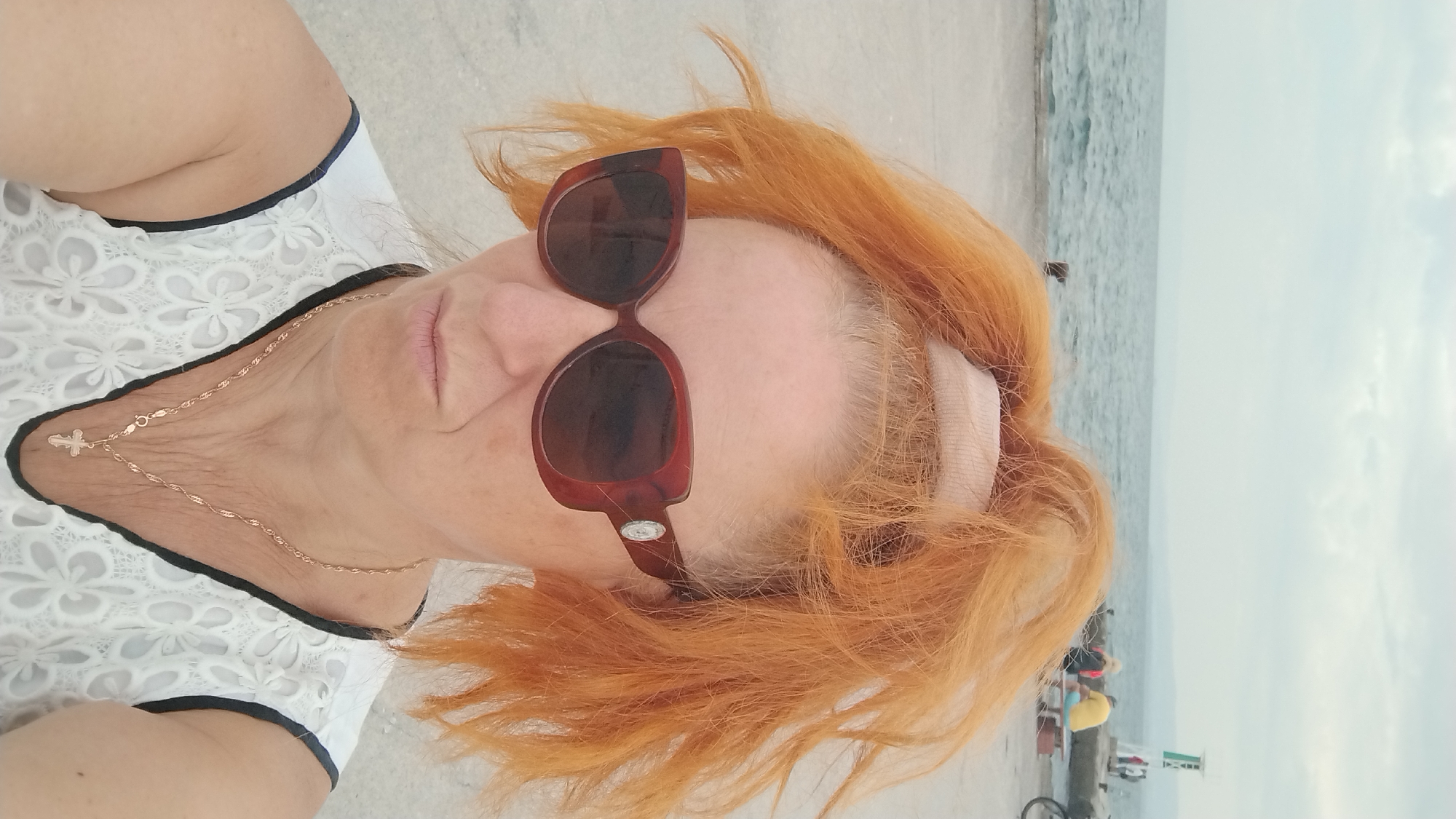 A medium shot selfie features a woman with bright orange, messy hair pulled back with a light pink band, wearing large, dark brown, butterfly-shaped sunglasses and a white sleeveless top with black trim and a floral lace overlay. She is also wearing a delicate gold chain with a small cross pendant. The woman is looking slightly to her left with a neutral expression. Behind her, the sea is visible with a pale sky overhead. In the background, there is a concrete pier with several people seated on a bench and a small lighthouse-like structure. The ground in the foreground appears to be a light-colored, sandy or paved surface. The scene is likely outdoors, possibly at a seaside location in Neoi Epivates, Greece, given the background elements.