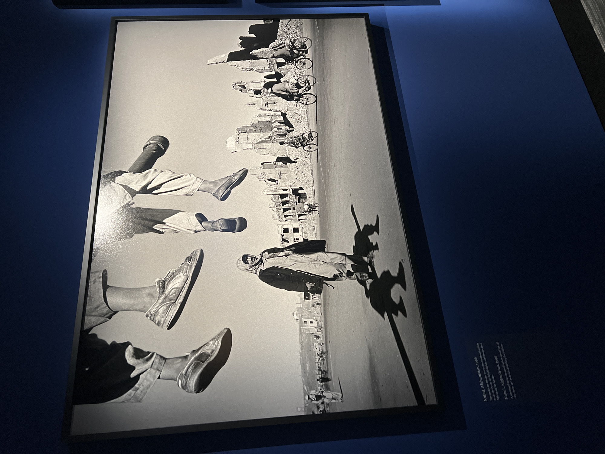 The image depicts a black and white photograph titled "Kabul, Afghanistan, 1996." It is framed and hanging on a dark blue wall. The photograph itself contains an urban scene featuring a figure standing on the ground casting a long shadow. In the background are bombed-out buildings. In the upper portion of the image, there are legs and feet appearing to be floating. In the upper portion of the image are bicycle riders. The photograph is displayed at a location in Berlin, Germany.