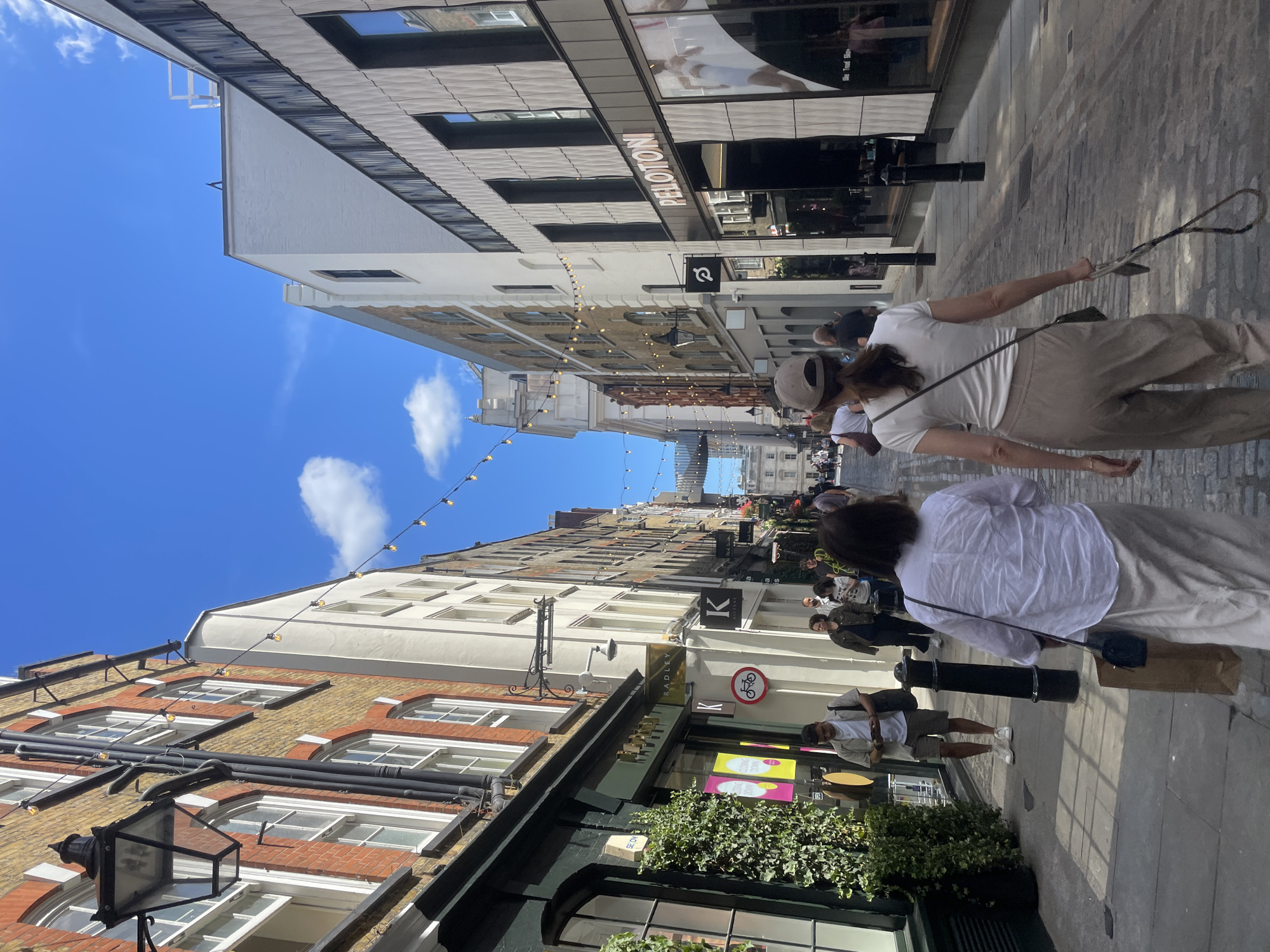 The image captures a street scene in London, United Kingdom. The perspective is from ground level, looking up and down a narrow, cobbled street lined with buildings. The day appears sunny with a bright blue sky and scattered clouds. String lights are suspended across the street between buildings, suggesting it might be an area known for its charm or events.
The scene is populated by several people walking or standing on the sidewalks. In the foreground, a woman in a white t-shirt and beige trousers is walking away from the camera, holding a strap in her hand. Behind her, another person, also in a white top and light-colored trousers, is walking. Further down the street, other individuals are visible, some browsing shop windows, others simply walking.
The buildings on either side of the street are a mix of architectural styles. Some have traditional brick facades with multiple windows, while others are more modern with large glass storefronts. Several businesses are identifiable by signage. The word "PELOTON" is prominently displayed on a building with a patterned facade. Another sign reads "RADLEY." A circular sign with a bicycle icon is also visible. The overall atmosphere seems to be that of a bustling shopping district on a pleasant day.