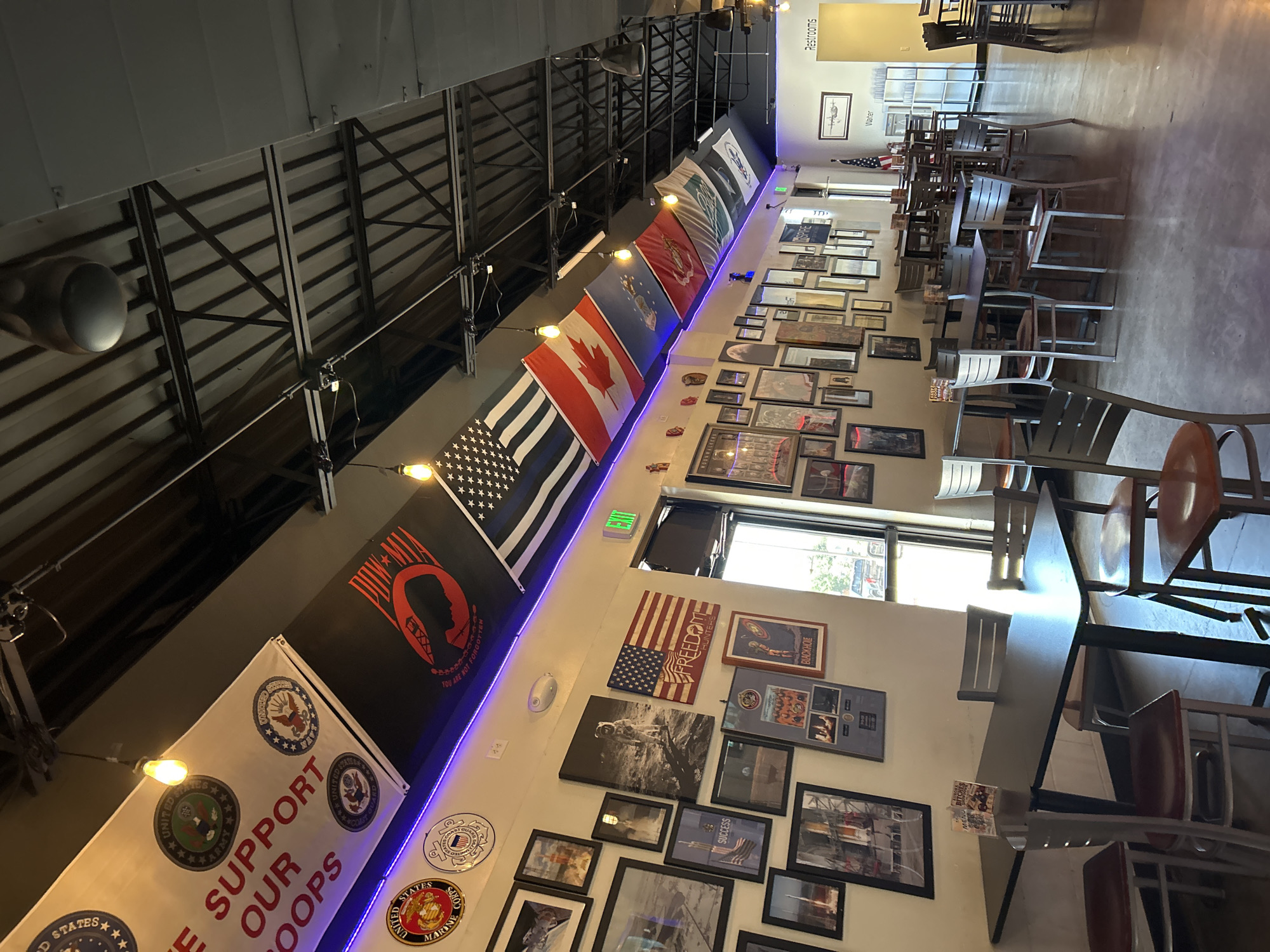 The image shows the interior of a restaurant, likely a sports bar or pub, decorated with military and patriotic themes. The walls are adorned with framed photographs and posters, many depicting space exploration and American flags. Various military service branch emblems and flags are displayed along the ceiling line, including the POW-MIA flag. The seating area consists of several tables with chairs, some of which are occupied by patrons. The lighting appears to be a mix of ambient and overhead fixtures. In the background, there is a visible "Restrooms" sign. The overall atmosphere suggests a casual dining establishment with a strong emphasis on military appreciation.