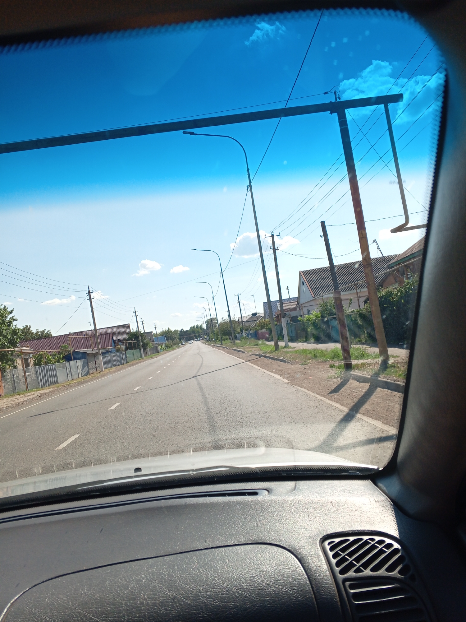 The image is taken from inside a vehicle, looking out through the windshield at a rural road scene in Novorozhdestvenskaya, Russia. The sky is bright blue with some scattered white clouds. A power line with multiple wires runs across the frame, supported by poles. To the right of the road, there are several houses with tiled roofs and fences, indicating a residential area. Trees are visible on the left side of the road. The road itself is asphalt with white lane markings. The dashboard of the vehicle is visible in the foreground, partially obscuring the view. The overall impression is a sunny day in a village or small town.