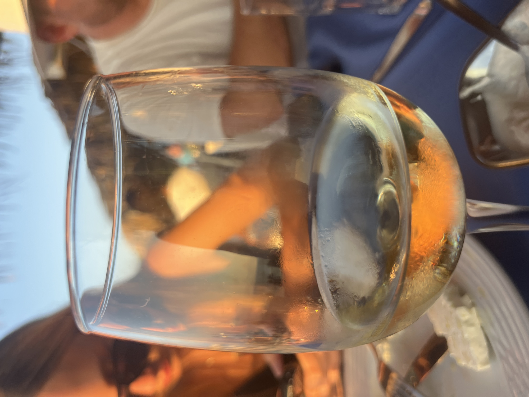 A close-up shot of a wine glass filled with white wine is positioned in the foreground. The glass is slightly condensation-covered. In the background, blurred figures of people are visible, suggesting an outdoor setting, possibly a restaurant or cafe, with warm, golden light indicating sunset or sunrise. The overall lighting is soft and warm, with a bokeh effect on the background elements. The context suggests a relaxed social gathering in Polis Chrysochous, Cyprus, likely at a seaside establishment given the ambient lighting and people enjoying a meal or drinks.