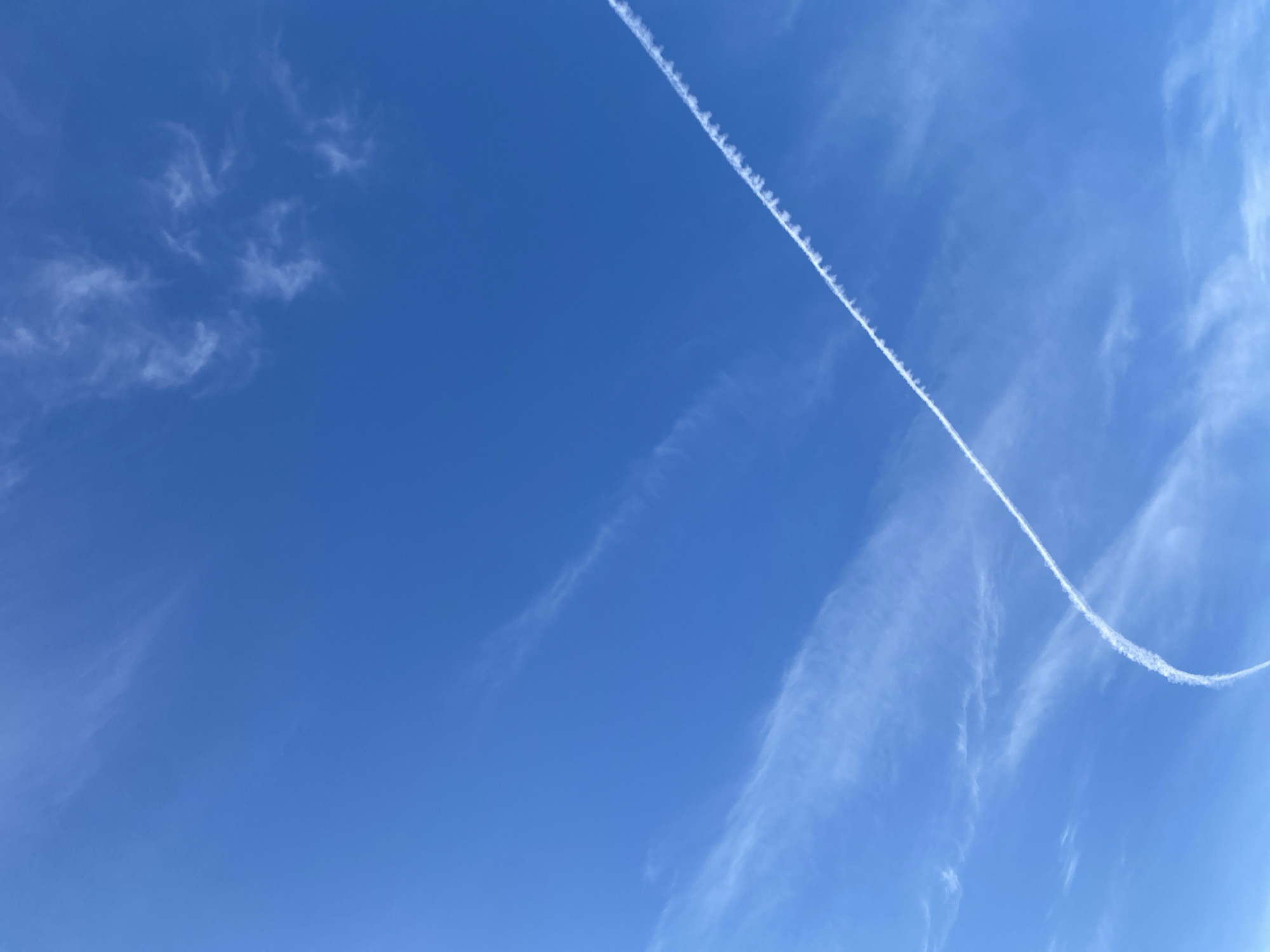 The image shows a bright blue sky with scattered wispy white clouds. A prominent white contrail, likely from an airplane, arcs across the sky from the lower left towards the upper right. The contrail is sharply defined in some areas and dissipates into fainter streaks of cloud in others. The overall scene depicts a clear, sunny day with evidence of recent air traffic over what is implied to be the area of Байқоныс ауылдық округі, Kazakhstan. There are no visible people or man-made structures in the frame.