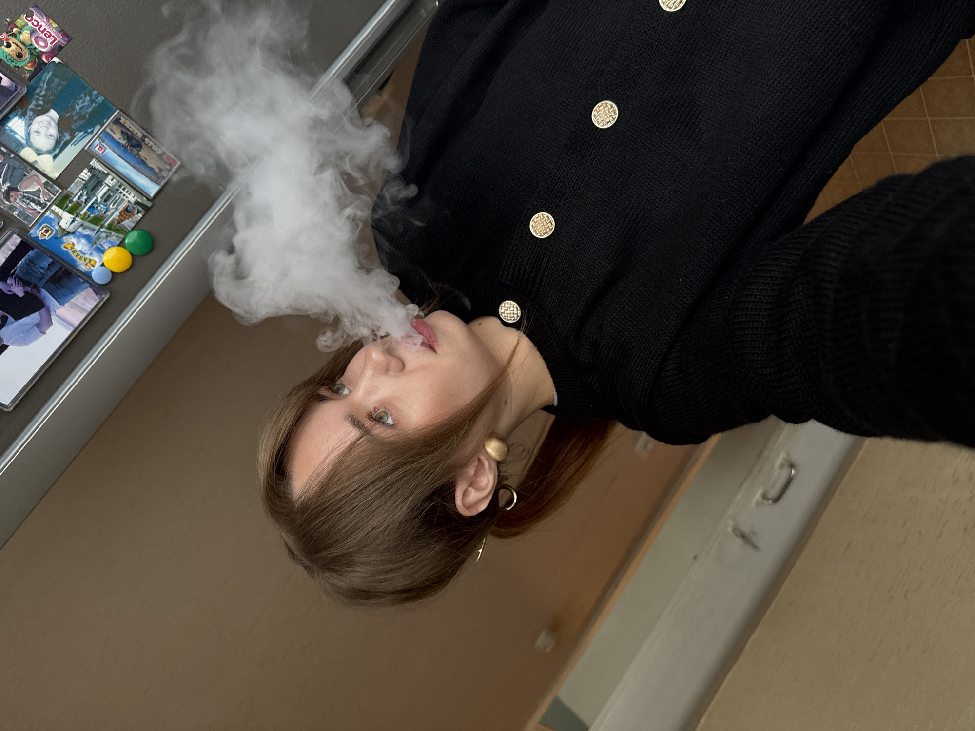 A woman exhales a cloud of white vapor into the air. She has light skin, green eyes, and brown hair partially tied back. She is wearing a black cardigan with gold buttons. The scene includes a refrigerator door adorned with magnets and a cabinet. The location is in the city of Chaikovsky, Russia.