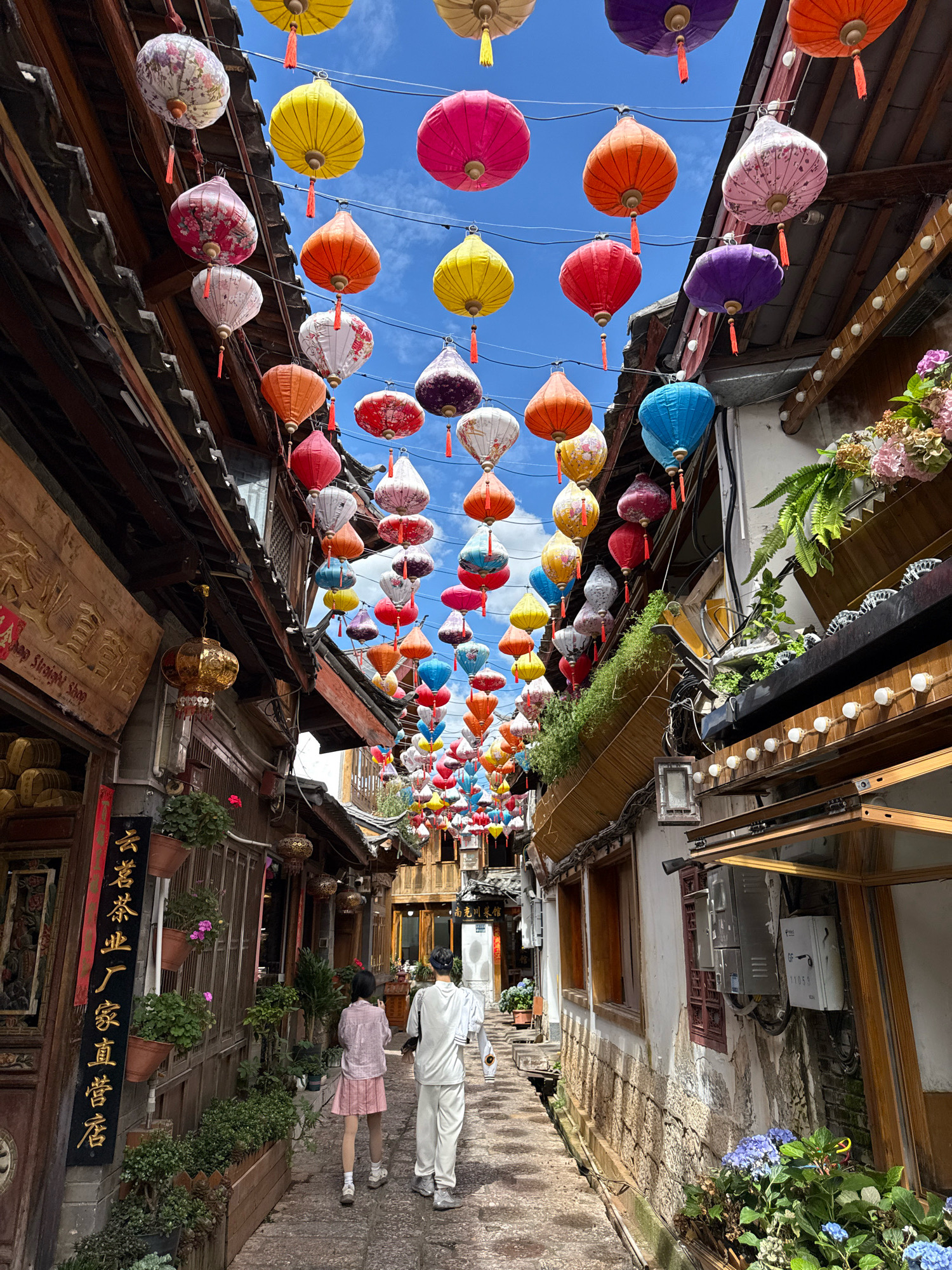The image depicts a street scene in Gucheng District, China, adorned with numerous colorful lanterns suspended overhead. The street is narrow, lined with traditional buildings featuring wooden eaves and textured facades. In the foreground, two individuals, a child and an adult, are walking away from the camera down the cobblestone path. The child is wearing a pink top and skirt, while the adult is in a white outfit. Shops with ornate signage and potted plants are visible along the sides of the street. The sky is blue with some clouds, suggesting a pleasant day. The lanterns, in various shades of red, yellow, pink, purple, and blue, create a vibrant canopy, typical of traditional Chinese street decorations.