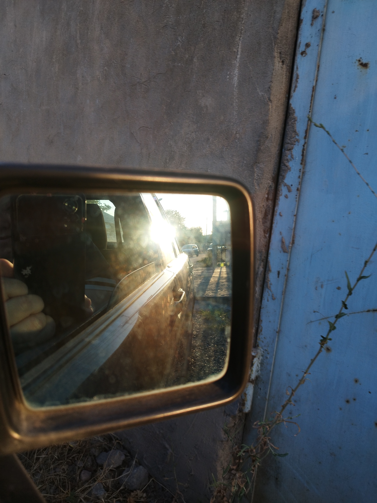 The image, taken on a sunny day, shows a close-up of a car's side mirror reflecting the surrounding environment and the interior of the car. The mirror is framed by a dark, slightly worn border. The reflection in the mirror displays a person's hand holding a dark-colored mobile phone, capturing the image. Also visible in the reflection is the car's interior, with seats and the sun shining brightly through the window, creating lens flare. Outside the car, the reflection shows a street with a few cars, trees, and a bright sky. The foreground of the image features the textured surface of a grey concrete wall to the left, and a weathered, light blue metal surface to the right. Dry grass and small rocks are visible at the bottom of the frame. The overall scene suggests an outdoor setting, possibly a parking area or roadside, in Shymkent, Kazakhstan, given the context.