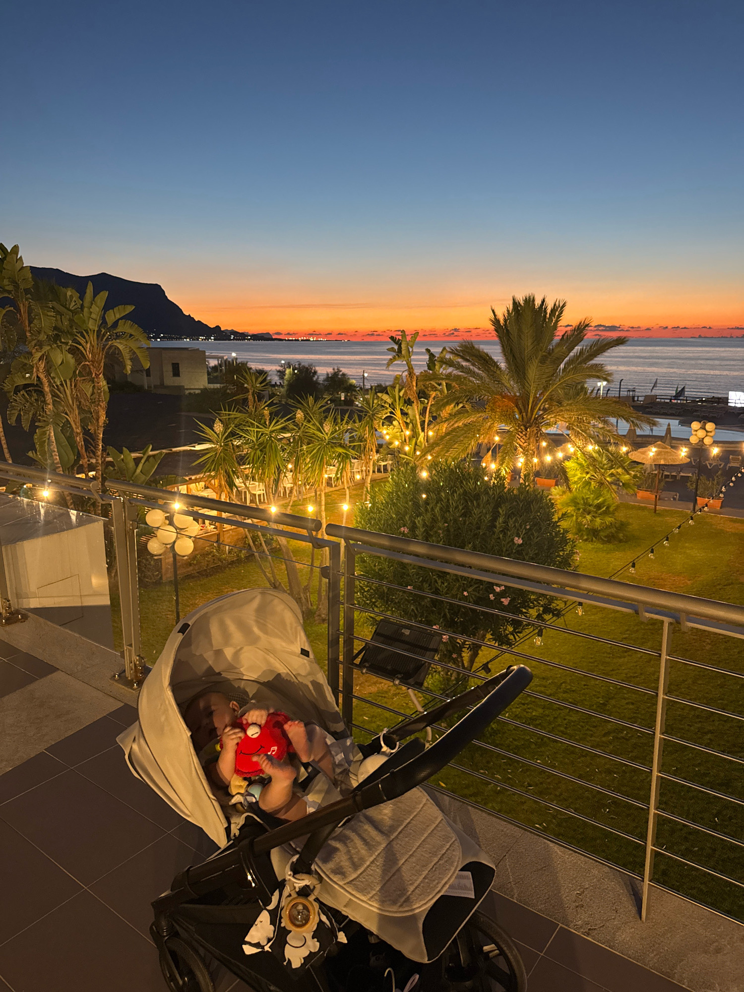 The image shows a baby sleeping in a stroller on a balcony overlooking a coastal town at dusk. The sky is a gradient of deep blue to orange, with the sun having just set. Palm trees and lush greenery dot the landscape, and string lights are visible throughout the scene, illuminating a resort-like setting. In the distance, a mountain range is silhouetted against the colorful sky. The stroller is positioned in the foreground, with the baby facing towards the viewer, holding a red toy. The overall atmosphere is serene and peaceful. The location is identified as Isola delle Femmine, Italy.