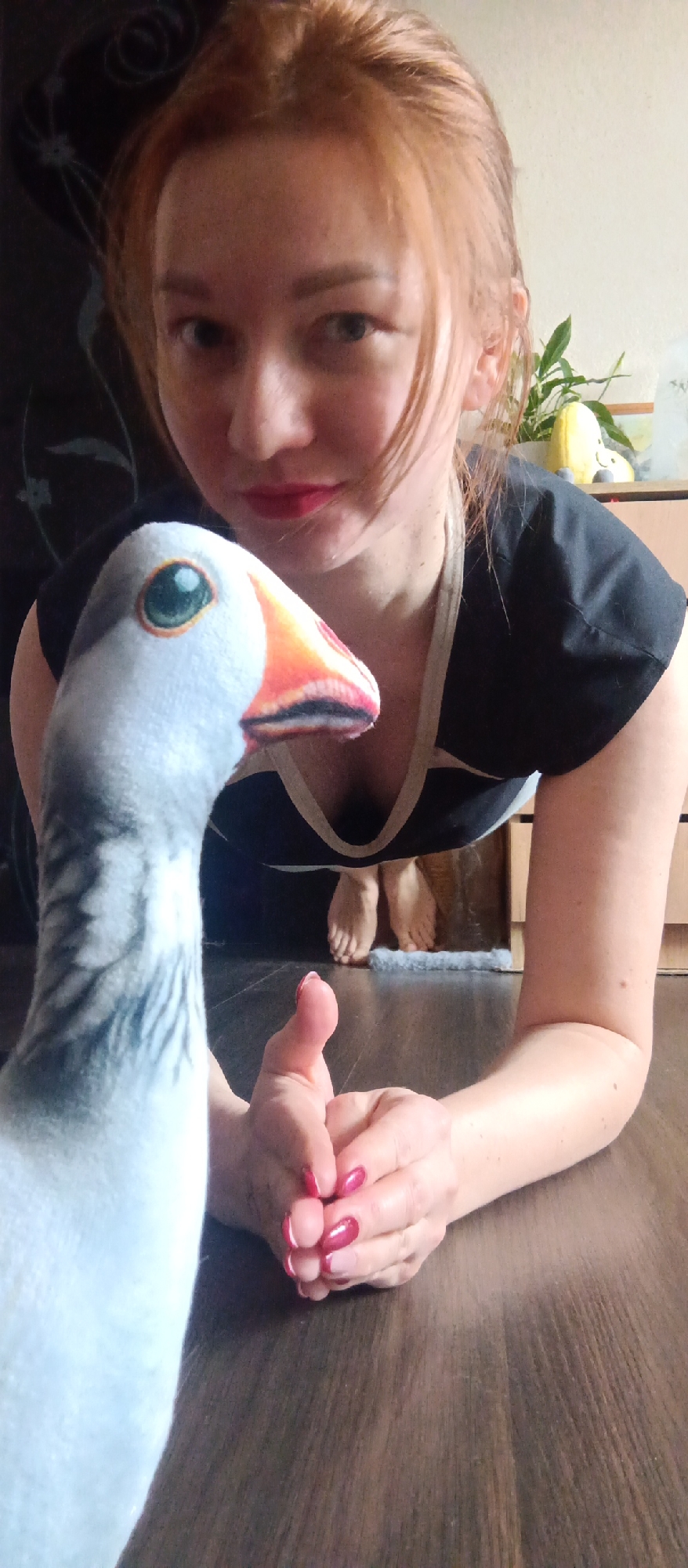 An indoor, eye-level shot features a woman and a plush goose toy. The woman, with reddish-blonde hair and a light complexion, is positioned low to the ground, appearing to be on her hands and knees or leaning forward. She wears a dark top with a light-colored trim and has red lipstick and bright red fingernail polish. Her expression is a subtle smile or smirk as she looks towards the camera.
In the immediate foreground, to the left, is a large plush goose toy. It has a white and grey body with a distinct green eye and an orange beak with black markings. The goose's head and part of its neck are prominent in the frame. The woman's hands, with their red nail polish, are clasped or resting on the dark wooden floor in front of her.
In the background, behind the woman, her bare feet are visible, resting on a small grey mat or rug. The room has a dark wooden floor. A dark wall with a subtle, dark pattern is visible behind her on the left, while a lighter wall and a light brown wooden cabinet or dresser are on the right. A green houseplant and a yellow, possibly moon-shaped, decorative item are placed on the cabinet. The lighting suggests it is daytime. There is no visible text in the image.