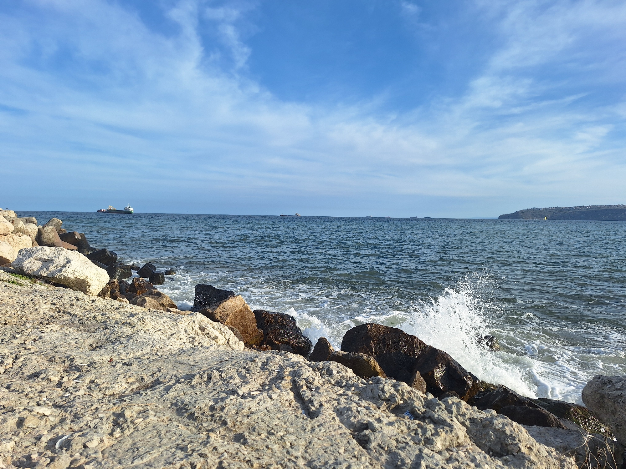 The image depicts a rocky shoreline in Varna, Bulgaria. Large, light-colored rocks and concrete form the foreground, transitioning to darker, wet rocks at the water's edge. Waves are crashing against the rocks, creating white splashes and foam. The Black Sea extends into the midground with several ships visible on the horizon. The sky is blue with scattered, wispy clouds. In the distance, the silhouette of the Asparuhovo neighborhood is visible on the southern edge of Varna Bay.
