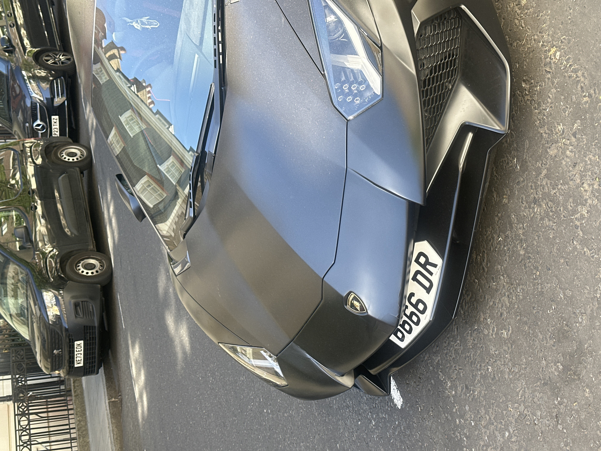 The image captures the front section of a dark grey, matte-finished Lamborghini supercar, parked on an asphalt street. The car's angular design, prominent headlights with LED arrays, and honeycomb grille are clearly visible. A small Lamborghini emblem is affixed to the front bonnet. The vehicle displays a British license plate, "G666 DR," mounted on its lower front.
The scene depicts a sunny daytime environment, with strong reflections of surrounding buildings, featuring multiple windows and architectural details, visible on the Lamborghini's windshield and bonnet. These reflections suggest a clear sky.
Other vehicles are parked nearby. To the left of the Lamborghini, a dark Mercedes-Benz car is partially visible, showing the license plate "P23 TKZ." Further back and to the left, a dark van or utility vehicle, possibly a Peugeot (indicated by a logo on its grille), is parked next to a black wrought-iron fence. This van bears the license plate "KE73 EOK."
The street surface is visible, along with a curb and a section of pavement. Shadows are cast across the ground, consistent with bright sunlight. No people are present in the image. The setting appears to be an urban street in London, United Kingdom, characterized by parked cars and architectural reflections.