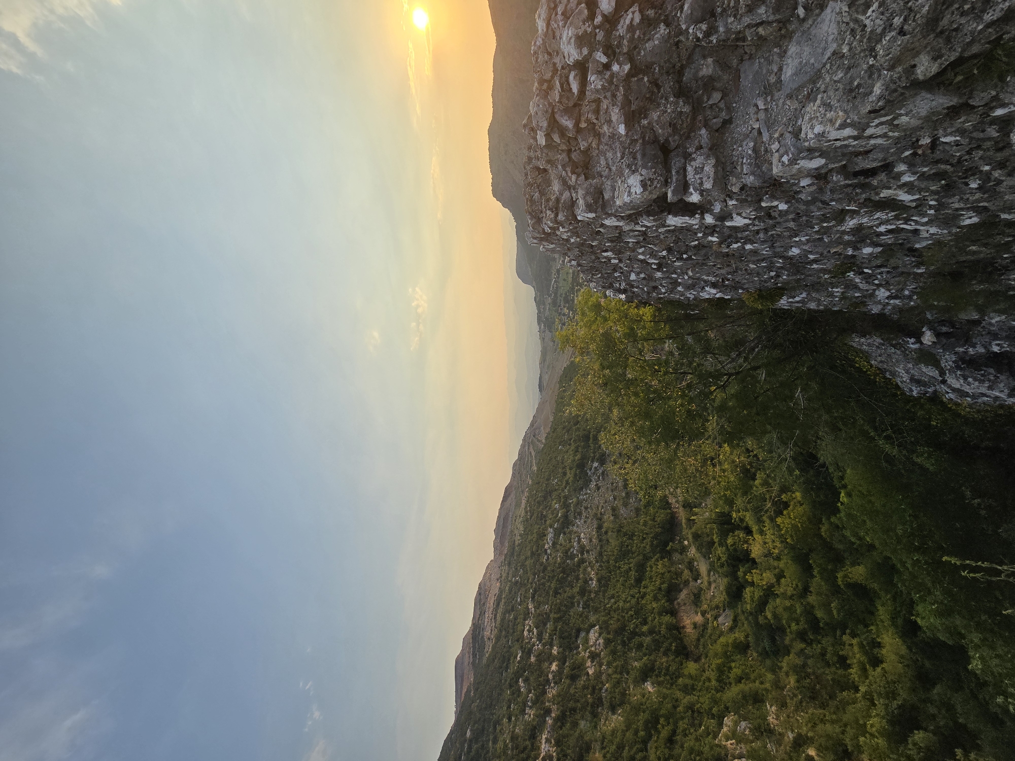 The image depicts a sunset view of the mountainous landscape surrounding Medun, Montenegro. In the foreground, to the right, a weathered stone wall made of rough-hewn rocks runs vertically, with some vegetation growing amongst the stones. To the left of the wall, lush green trees and dense foliage cover the side of a steep, rocky hill.

Further into the distance, the landscape unfolds into rolling hills and mountains, blanketed in a mix of green vegetation and exposed rocky terrain. The sky above is a gradient of soft blues at the top, transitioning to warmer oranges and yellows near the horizon, indicating the setting sun. The sun itself is a bright, glowing orb positioned in the upper right quadrant of the sky, casting a warm light over the scene. Faint, wispy clouds are scattered across the sky. In the far distance, a hazy valley with scattered structures can be observed, suggesting a settlement. The overall impression is one of natural beauty and historical significance, hinting at the ancient fortress of Medun which is located in this area.