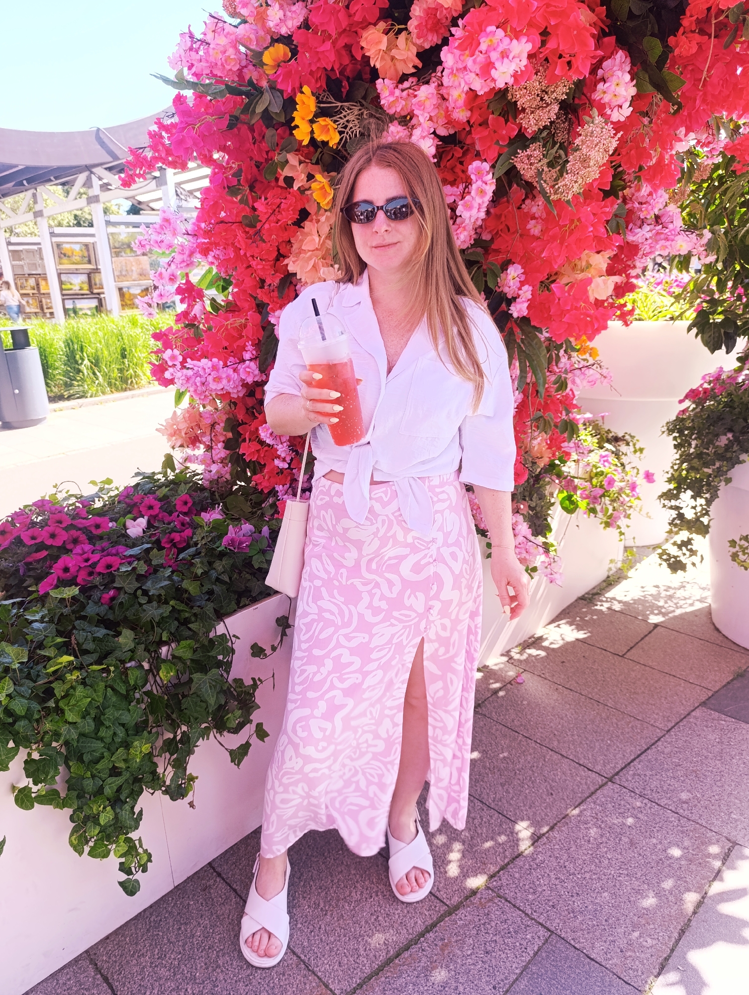 A young adult female stands outdoors, facing slightly left of the camera, in an unknown urban or park-like setting. She has long, reddish-blonde hair, wears dark sunglasses, a white collared shirt tied at the waist, a long pink and white patterned skirt with a high side slit, and white flat sandals. A small cream-colored shoulder bag hangs on her left side. She holds a clear plastic cup with a lid and black straw, containing a reddish-orange beverage, in her right hand, where pale yellow nail polish is visible. Her left hand hangs at her side, also showing pale yellow nail polish.
Behind her is a large, dense floral display featuring vibrant pink and red flowers, with some orange and yellow blooms intermingled, resembling bougainvillea. To her immediate left, a white planter box is filled with dark green ivy and small purple and pink petunia-like flowers. To her right, a wider white planter contains spilling greenery. The ground is paved with light grey rectangular stones, displaying dappled sunlight and shadows. In the mid-ground to the left, a metal pergola structure, green foliage, and a dark grey cylindrical trash receptacle are visible under a clear blue sky.
