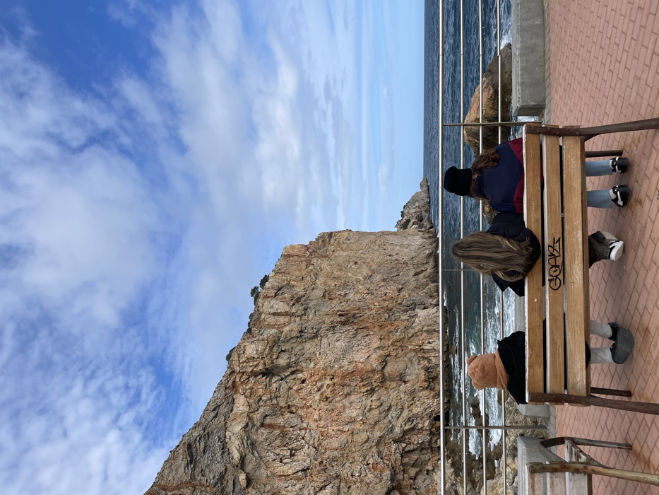 The image displays a coastal scene in Torroella de Montgrí, Spain, featuring three individuals seated on a wooden bench with their backs to the camera. The bench, which has metal supports, shows graffiti on its backrest. From left to right, the individuals are observed wearing a brown, possibly knitted, hat and dark jacket; a dark jacket revealing long, light brown hair; and a black beanie with a multi-colored dark blue and red jacket. They are positioned on a red-brown paved promenade, facing the Mediterranean Sea, which features dark blue water and white breaking waves against rocks and the base of a large cliff. A metal railing separates the paved area from the water. A significant, rugged reddish-brown rock cliff dominates the left side and background, exhibiting visible geological strata and sparse vegetation atop its summit. The sky is partially cloudy with scattered white clouds against a blue background. A fragment of another wooden bench or table is visible to the far left.