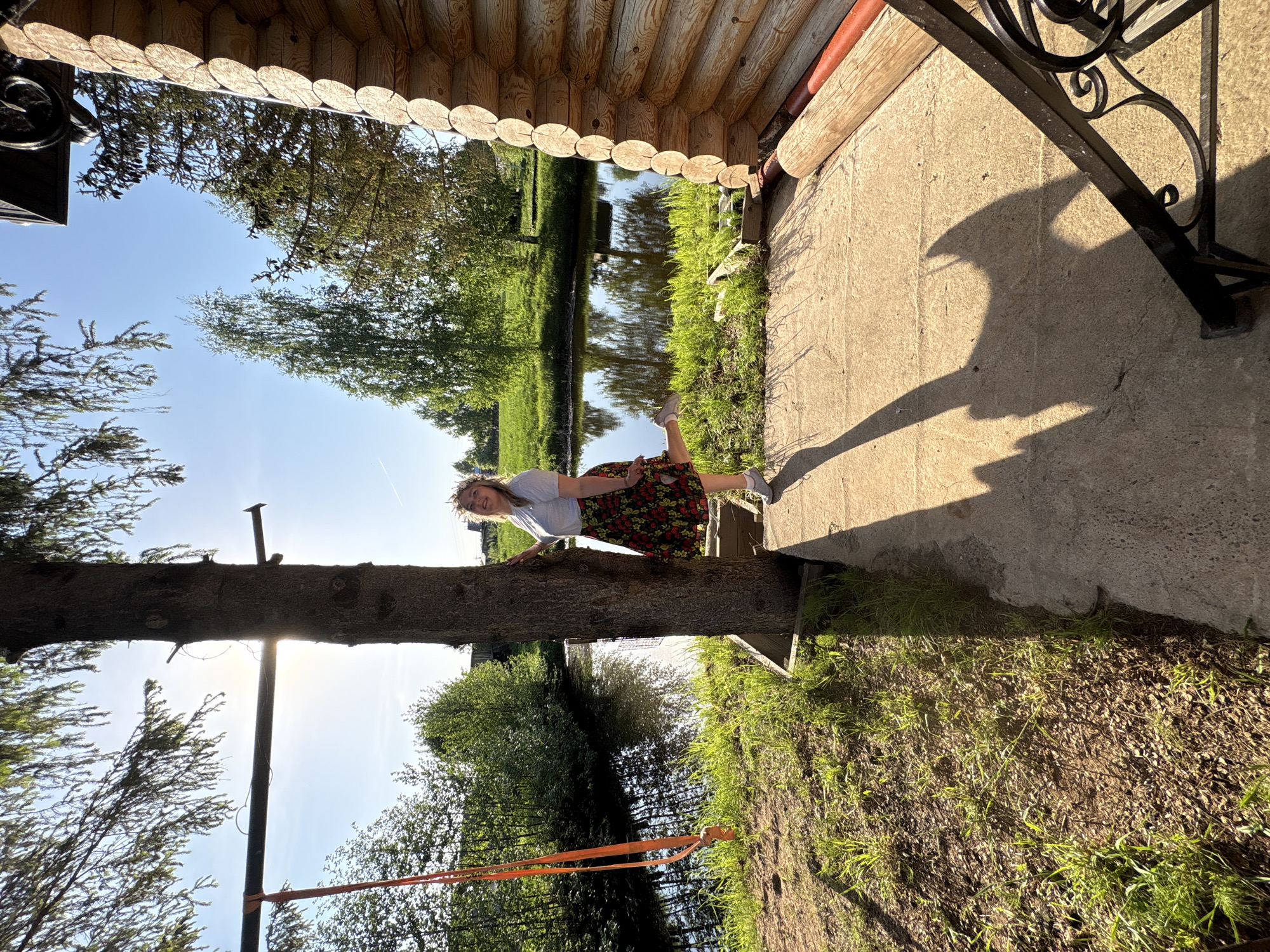 A woman is balanced on a horizontal tree trunk in a backyard setting. The trunk is supported by a vertical post and has an orange strap attached. The woman is wearing a white shirt and floral skirt, smiling toward the camera. The location appears to be near a body of water. The image is taken from under an overhanging structure that provides a partial view of a wooden cabin. The shadow of wrought iron details is cast on the ground surface near the cabin. The location is in Markovo, Russia.
