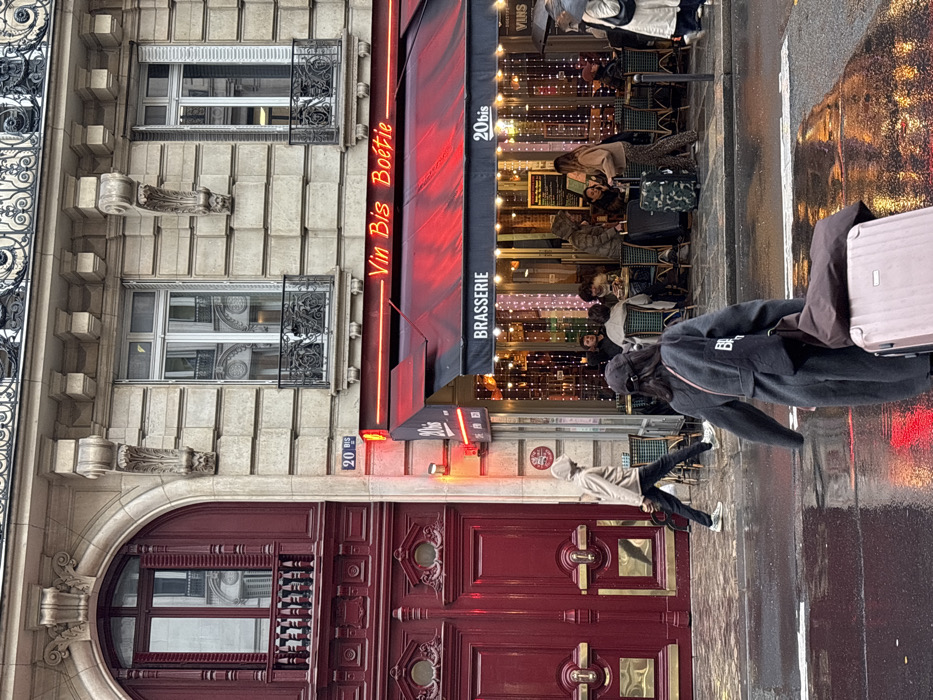 The scene depicts a wet street in Paris, France, specifically at **20 bis Rue La Boétie**, indicated by visible street numbering and brasserie signage. The image captures an evening or nighttime setting, with artificial light illuminating the scene.
On the right, a brasserie named "Vin Bis Boëtie," also identified as "20 bis," features a black awning with "BRASSERIE" printed on it. Above the awning, a red neon sign spells out "Vin Bis Boëtie." The brasserie's interior is brightly lit with numerous vertical string lights, showcasing multiple patrons seated at small tables, engaged in activity.
To the left of the brasserie, an ornate, deep red wooden door with intricate carvings and gold accents dominates the ground floor of an adjacent building section. Above this door is a matching arched window with open shutters. The upper floors of both structures exhibit classic Parisian Haussmannian architecture, characterized by light-colored stone, decorative window sills, and wrought-iron balconies.
The cobblestone street and sidewalk are visibly wet, reflecting the red neon glow from the brasserie. In the immediate foreground, a person, seen from behind, wears a dark coat and baseball cap, pulling a rose gold wheeled suitcase across the wet street, moving away from the camera. Further back on the sidewalk, in front of the red door, another individual wearing a light-colored hooded jacket and dark pants walks from right to left. On the far right, partially obscured, a person holding an open umbrella is visible on the sidewalk. Near the brasserie entrance, a fourth individual stands beside a camouflage-patterned wheeled suitcase.