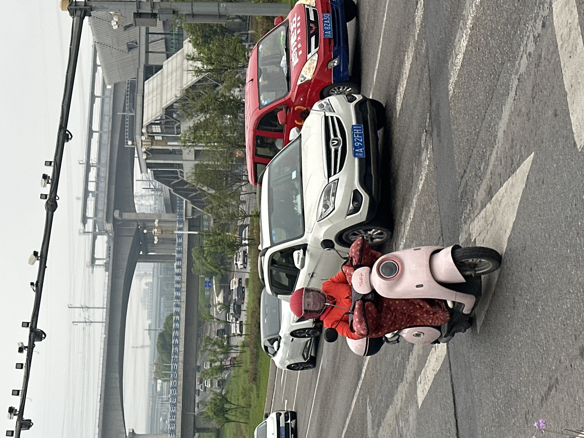 This image, taken in Jiangjin, China, captures a street scene with a person on an electric scooter and several vehicles. The photograph is oriented vertically, showing the street with traffic moving and a large, multi-level infrastructure in the background.
In the foreground, a person wearing a bright orange-red jacket and a matching red helmet is seated on a light pink electric scooter. The scooter has a red and white patterned cover over its handlebars and seat, and the word "Vintago" is visible on its front panel. The person appears to be stationary or moving slowly in a lane of traffic.
Immediately behind the scooter is a white SUV, identified by its blue license plate "渝 A 92FH1". Chongqing's (渝, Yú) abbreviation is seen on the plate. Behind the SUV is a red commercial van, possibly a delivery vehicle, with the blue license plate "渝 A 8ZA30". The Chinese characters "百世快递" (Best Express) are visible on the van's front windshield. Other vehicles are present further along the road.
The road surface is asphalt with white lane markings and arrow indicators. To the far left, a tall utility pole equipped with multiple surveillance cameras is prominent. The background is dominated by a vast concrete bridge or overpass structure with multiple spans and supports, suggesting a significant transportation artery, possibly a railway or elevated highway. The sky is overcast or hazy, indicating daytime without direct sunlight. Green foliage lines the roadside and is visible beneath the bridge structure.