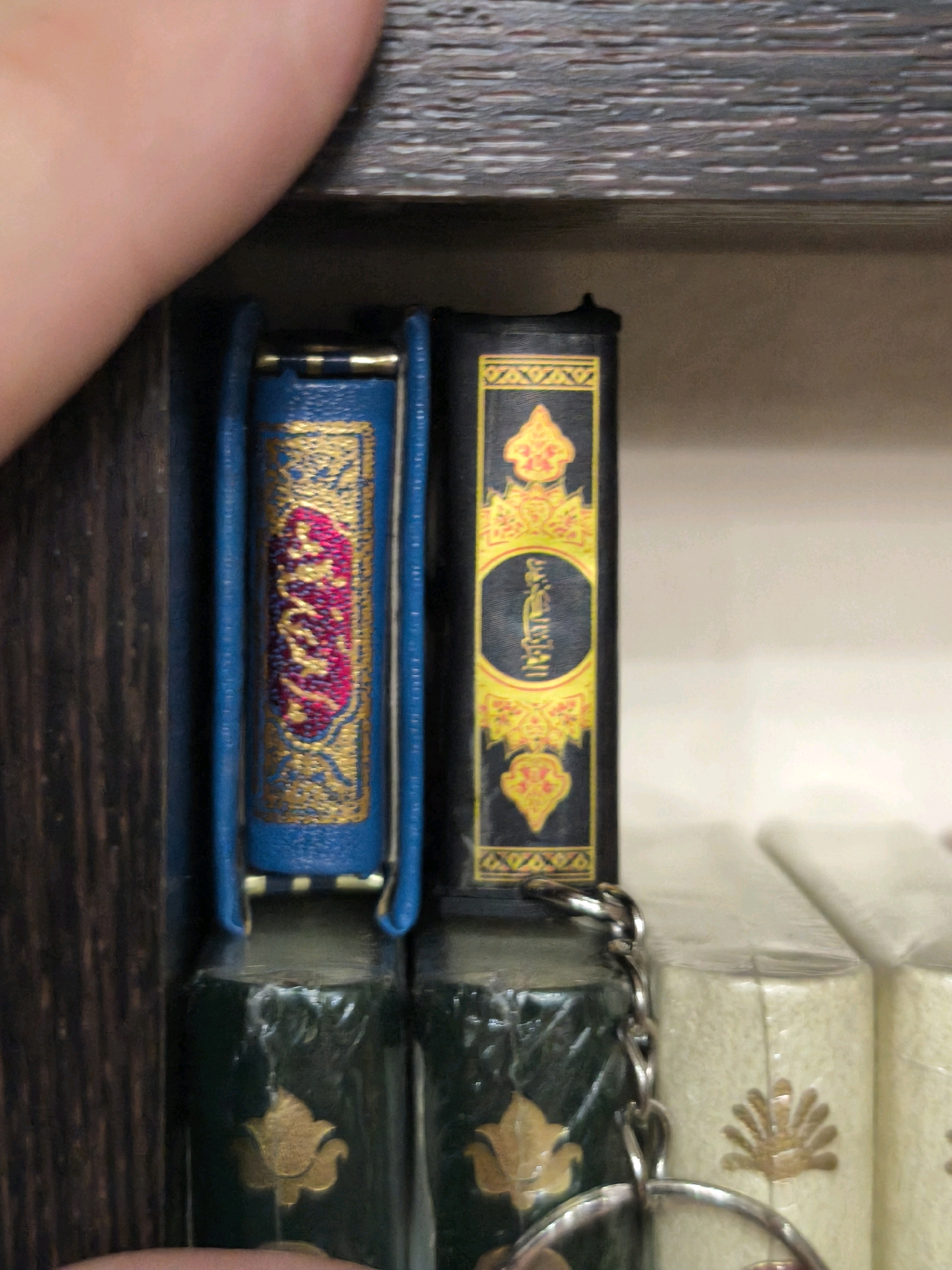 This is a close-up, eye-level, indoor shot of two miniature books displayed on a wooden shelf. The books are standing upright, with the spine of the left book facing the viewer. The left book has a blue cover with intricate gold ornamentation and pink text. The right book has a black cover with a circular emblem containing yellow text and decorative gold patterns at the top and bottom. Both books appear to be part of a larger collection, as suggested by the edge of another book visible to the right, covered in cellophane and featuring gold embossed patterns. A silver chain with a circular ring is attached to the top of the right book, suggesting it may be a keychain. The background is blurred, showing a textured wooden shelf above and a plain wall or surface behind. The image is well-lit, with focus primarily on the two miniature books. The specific location within Karachayevsk, Russia, is not discernible from the visual information provided in the image.