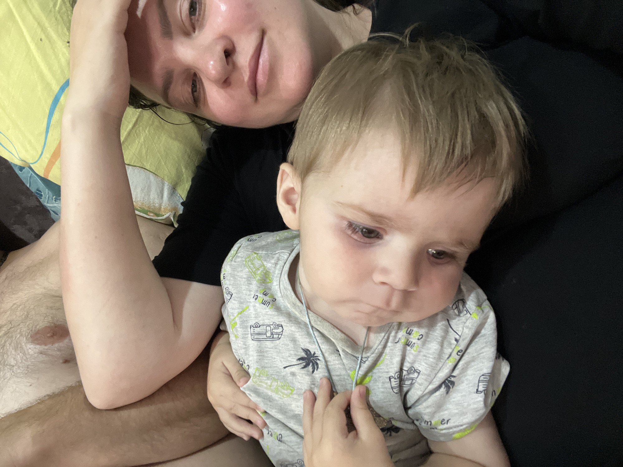 An adult's arm is resting on a pillow, with the adult's face visible in the upper left of the frame, looking upwards. To the right of the adult, a toddler is seated, looking downwards with a furrowed brow. The toddler is wearing a grey t-shirt with a pattern of palm trees and cars. The background consists of dark fabric. The scene appears to be indoors, possibly in a bedroom, though no specific location within Noviy, Russia, can be determined from the image.
