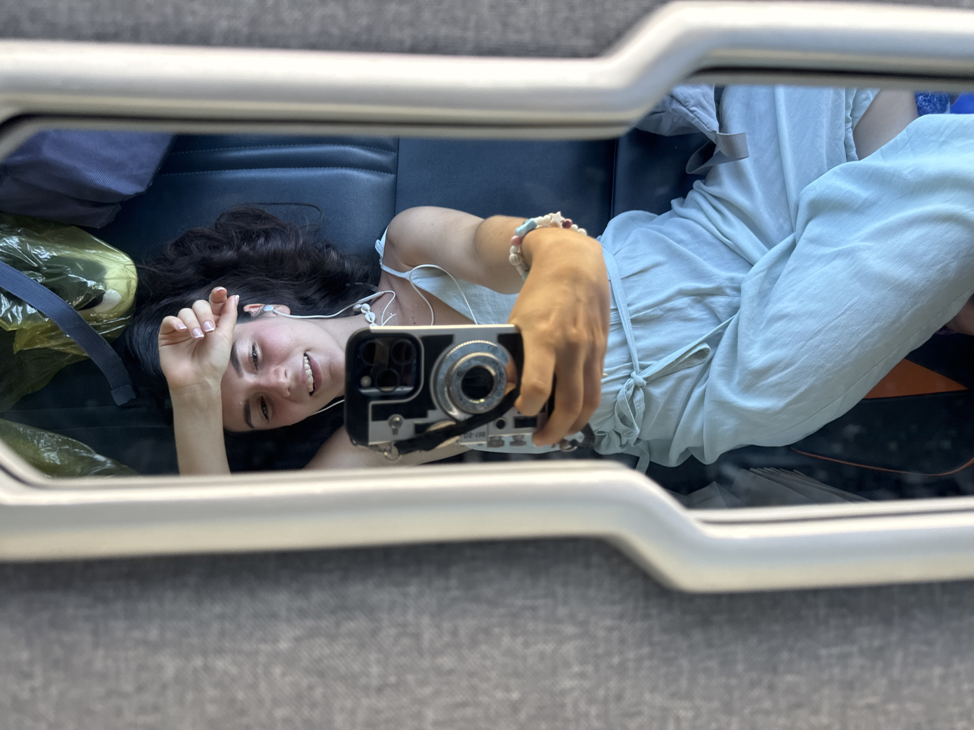 A young woman is lying down taking a selfie in a mirror, presumably on a bus or train. She has dark curly hair, is wearing a light-colored top and pants, and has earphones in. She's smiling. There is a backpack and a plastic bag next to her. The reflection shows what appears to be a bus or train seat. There is no information available to confirm the location.