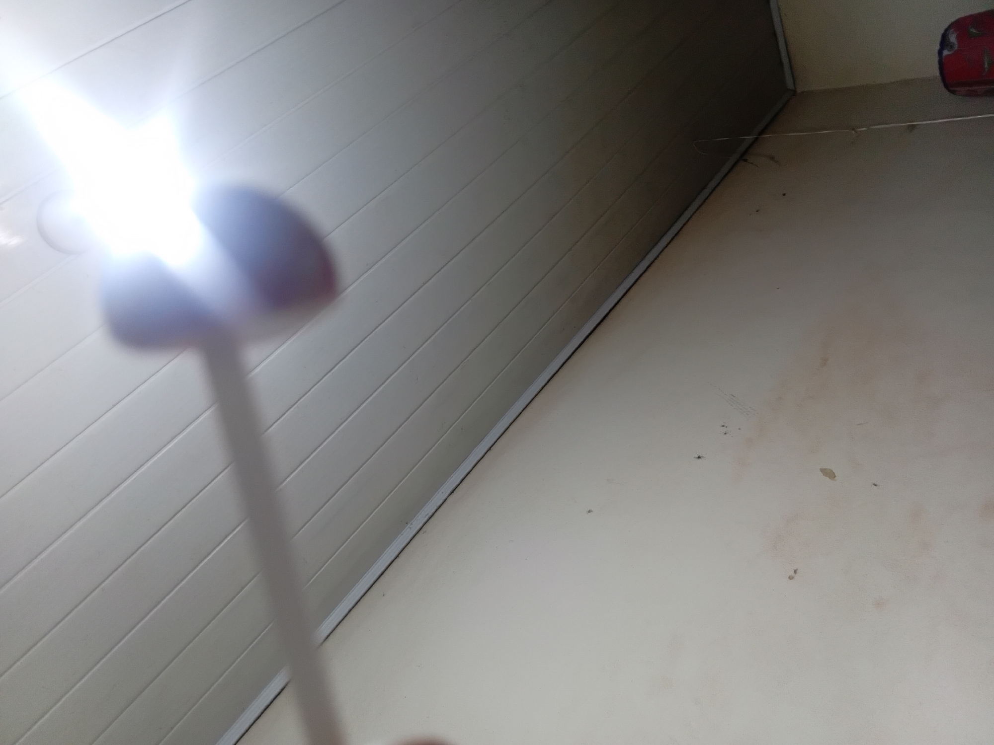 A bright light source, likely a flashlight, is angled towards a white, slatted ceiling, creating lens flare and a blurry purple object in the foreground. The wall below the ceiling is a pale beige color with visible water stains and dark marks. The scene appears to be indoors, possibly in a room or hallway in Ejule, Nigeria. The lighting suggests the image was taken during the day or with artificial light in a dimly lit environment.