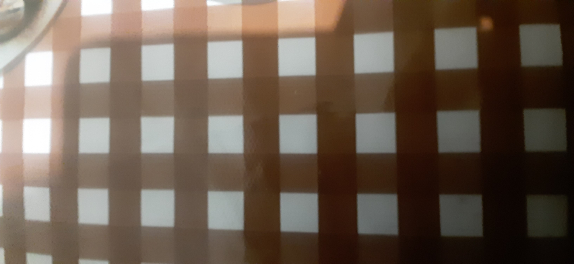 The image shows a close-up, abstract view of a brown and white checkered pattern. The pattern is consistent and appears to be a surface like a tablecloth or a printed material. There are no people, objects, or specific environmental details visible that would allow for identification of a location within Yessentukskaya, Russia or any other city. The lighting suggests it is indoors or in an area with artificial light, casting some shadows or highlights on the surface.