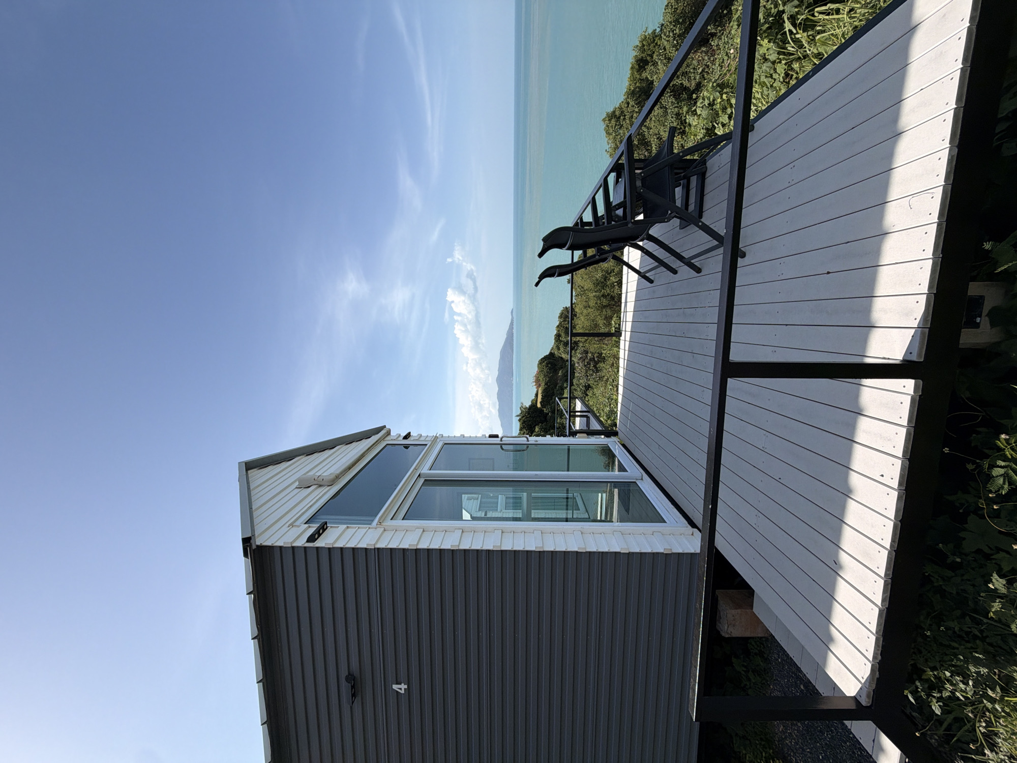 A modern, minimalist cabin with a grey corrugated metal exterior and white wood siding stands on stilts overlooking a turquoise ocean. The cabin features a large glass sliding door and a rectangular window above it, allowing for expansive views. A light-colored wooden deck extends from the cabin, furnished with two black folding chairs facing the sea. The deck is bordered by a black metal railing, and lush green foliage surrounds the structure, suggesting a natural, somewhat secluded location. In the distance, a hazy island or landmass is visible on the horizon under a bright blue sky with scattered white clouds. The scene evokes a sense of tranquility and a holiday retreat, likely situated in a coastal area of Ko Samui District, Thailand, known for its picturesque beaches and ocean vistas.