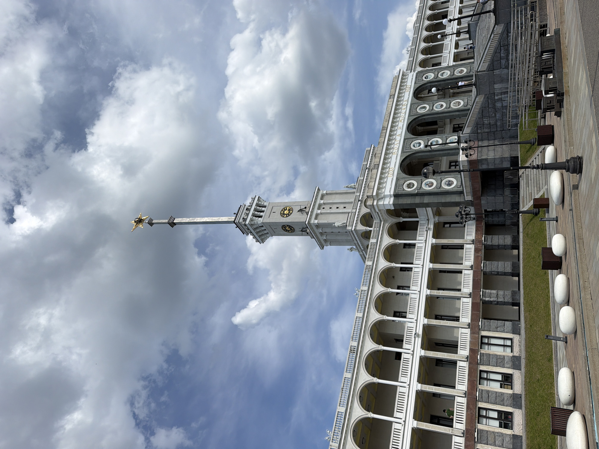 The image depicts the Moscow Canal Administration building in Khimki, Russia, viewed from a low angle. The building has a multi-story architectural design with white columns and arches. A tall tower with a clock is visible. A spire topped with a star extends into the sky. The facade of the building reads "VOLGA MOSKVA 1937," referring to the construction date of the Volga-Moscow Canal. A paved area with streetlights and seating is visible in the foreground. The sky contains white clouds against a blue backdrop.