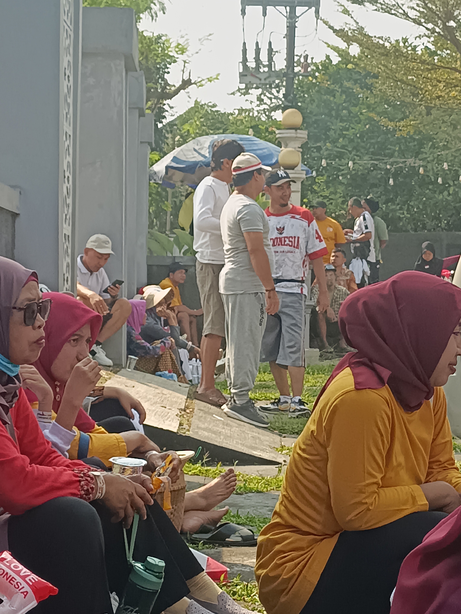 In Sleman, Indonesia, a group of people are gathered outdoors. In the foreground, several individuals are seated. To the left, a woman wearing a red shirt and black pants, with a dark headscarf and sunglasses, sits with her hands in her lap. Next to her, a woman in a red headscarf is also seated. In the center of the frame, a man wearing a white shirt and beige cap is seated and looking at his phone. Behind him, a man in a grey t-shirt and grey shorts, along with another man in a red and white shirt with "INDONESIA" printed on it, are standing. Other individuals are visible in the background, some standing and some seated, amidst greenery and structures that appear to be part of an outdoor public space. The lighting suggests it is daytime, with sunlight casting shadows.