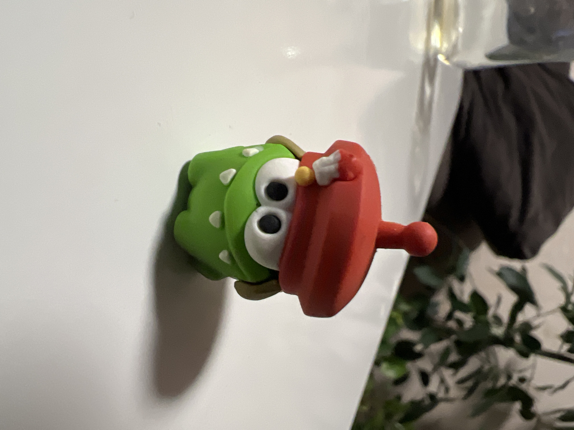 A green toy figure with large white eyes and tiny white teeth is positioned on a white surface. The figure is wearing a red beret with a white tuft and a small yellow embellishment. In the background, a blurred glass and dark fabric can be seen, along with out-of-focus green foliage. There is no indication that this scene is located in Yekaterinburg, Russia.