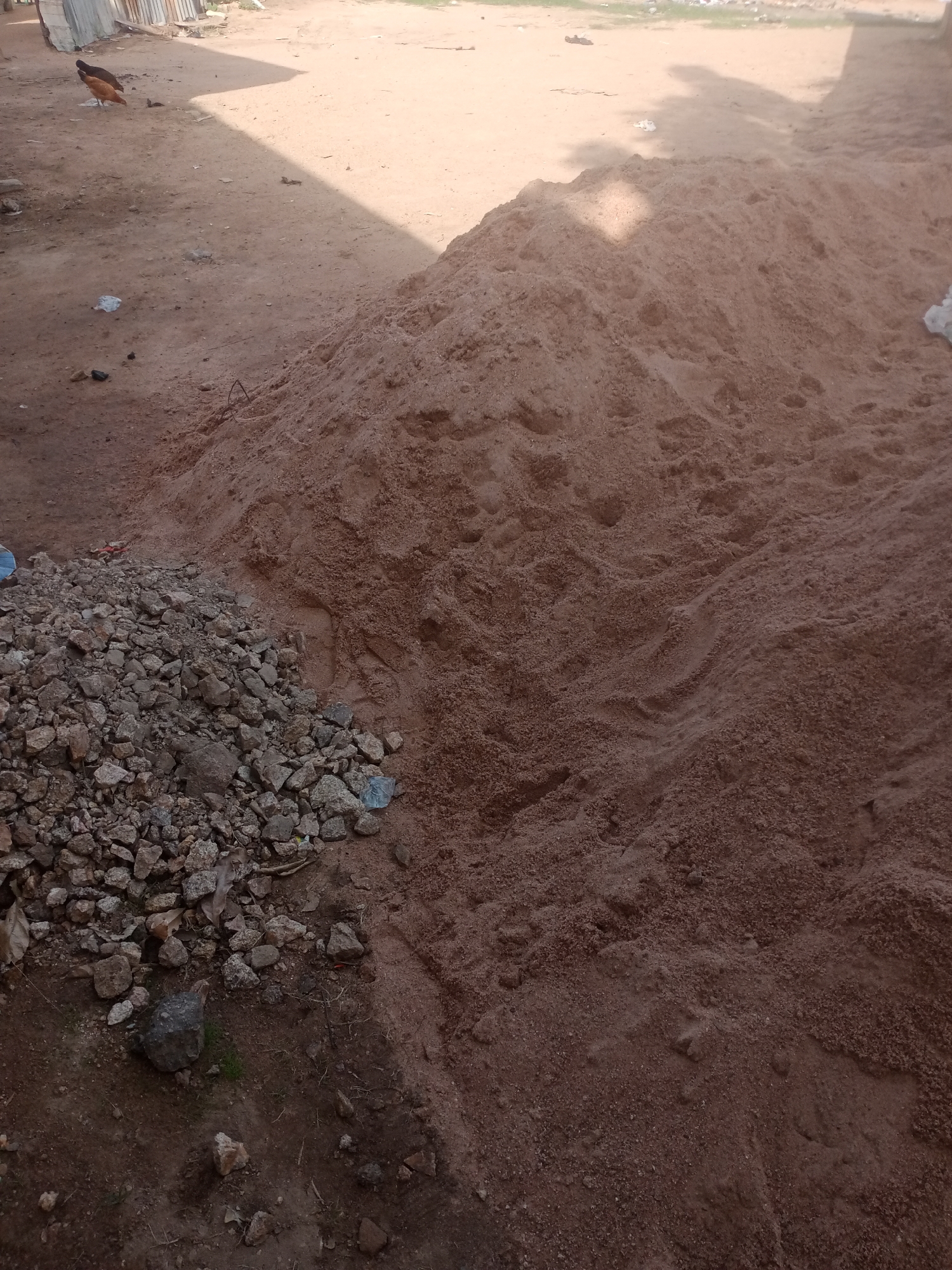 In Jalingo, Nigeria, a pile of sand is situated on a dusty, unpaved ground. To the left of the sand pile, a smaller heap of rocks and gravel is visible. In the background, the ground continues to be dusty and uneven, with some scattered debris. A shadow falls across the scene from an unseen structure to the left, casting a diagonal line over the sand and ground. Two chickens, one brown and one black, are pecking at the ground in the distance, near a corrugated metal fence. The overall environment appears to be an outdoor, undeveloped area.